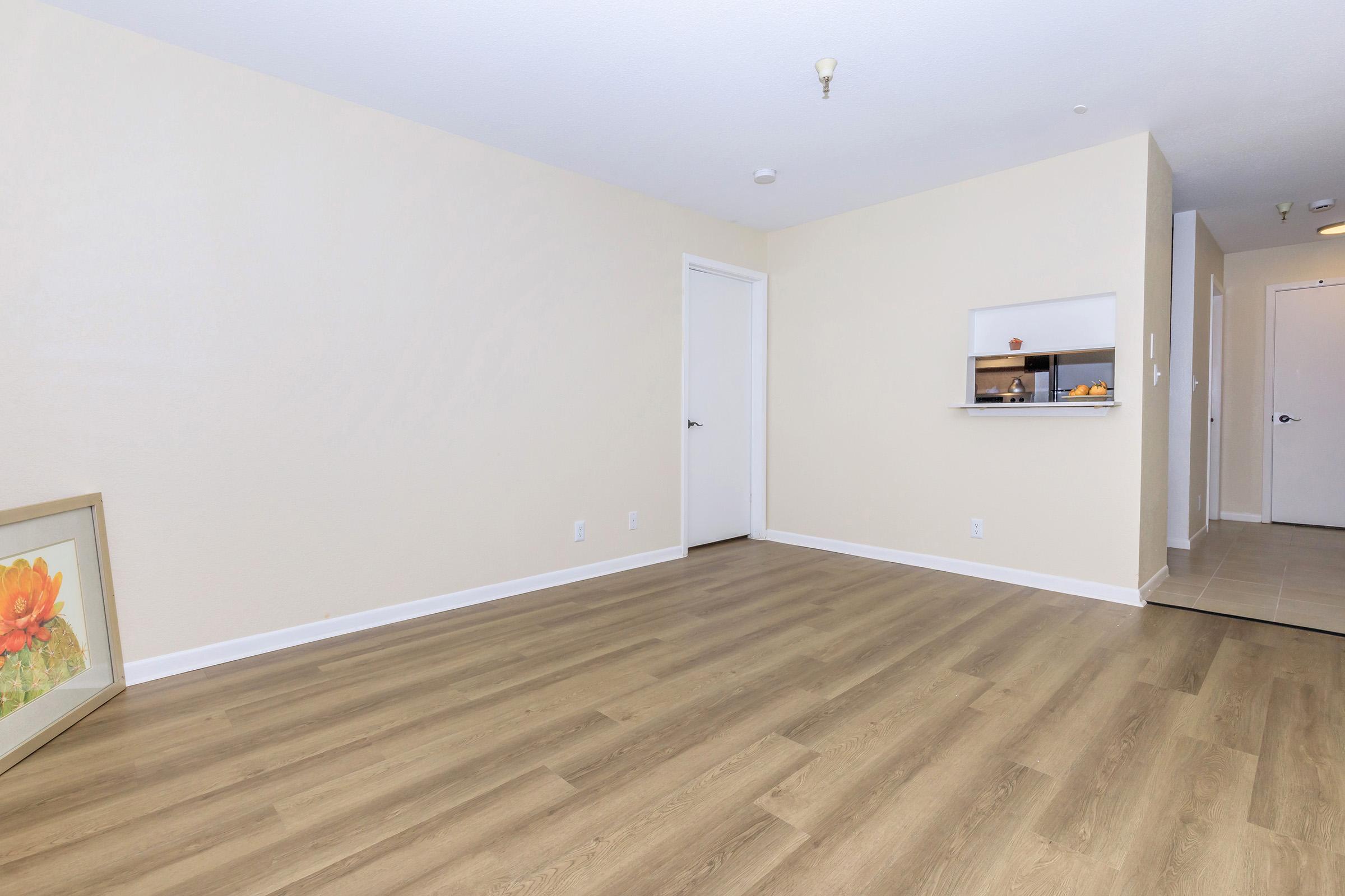 A spacious, empty room with light-colored walls and wooden flooring. There is a small opening with a counter leading to a kitchen area. A door is visible on the left, and another hallway extends to the right. A framed floral artwork is placed on the floor near the wall.