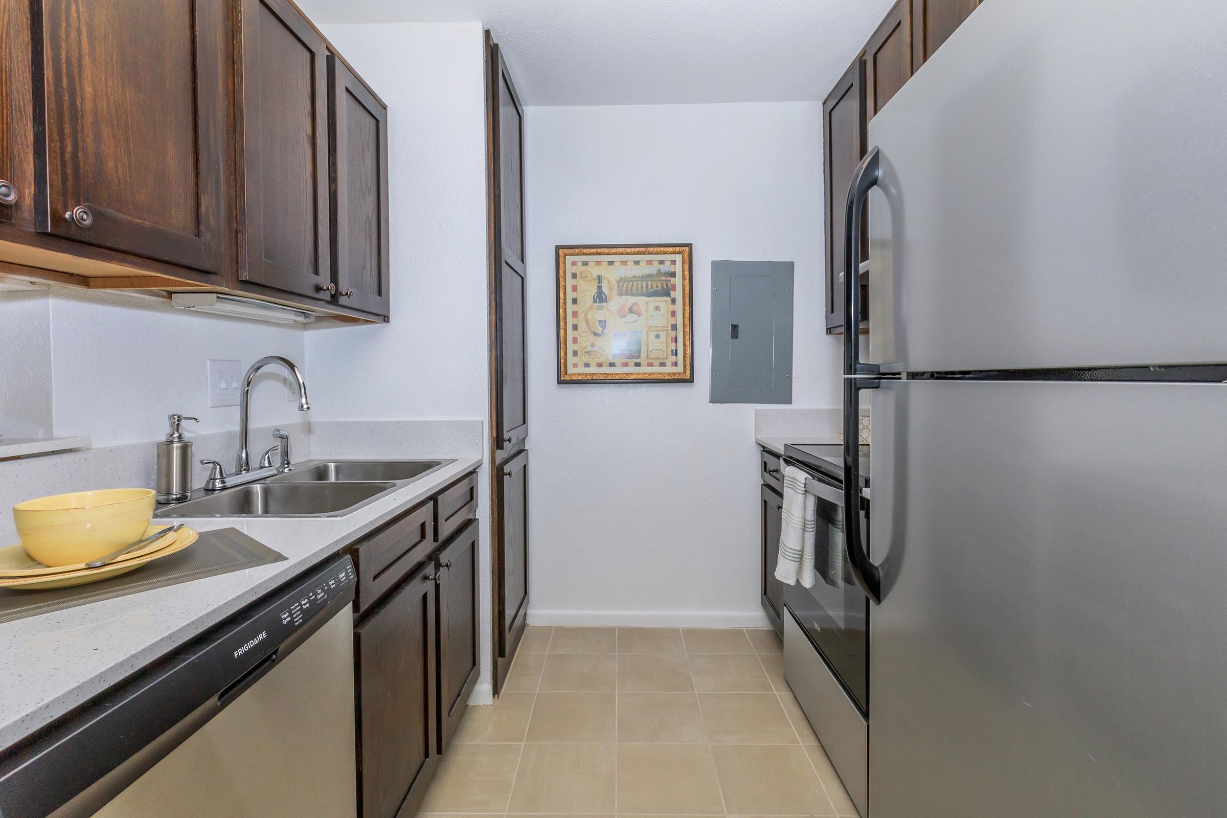 A modern kitchen featuring dark wood cabinets, stainless steel appliances including a refrigerator and dishwasher, a sink with a faucet, and a beige tiled floor. There is a decorative framed artwork on the wall, adding a touch of style to the space.