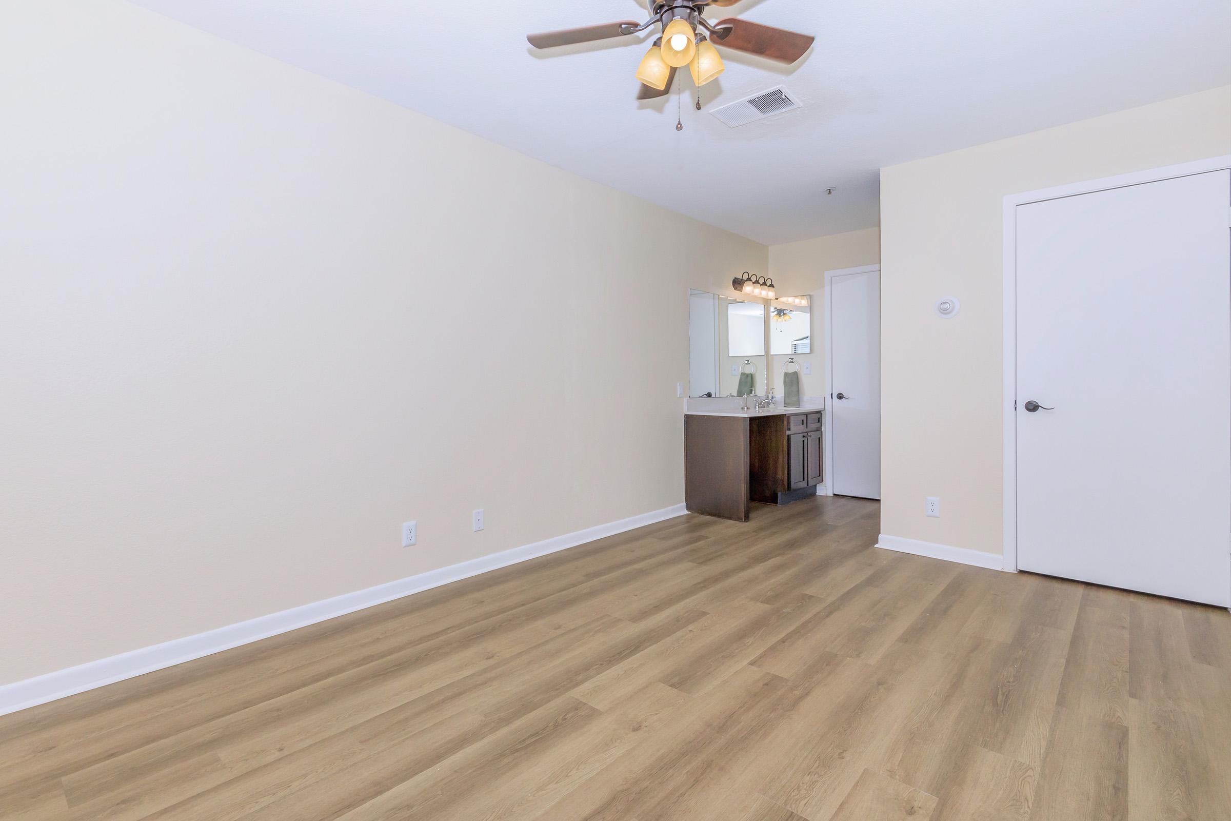 A spacious room with light beige walls and light wood flooring. A ceiling fan hangs above, and there is a bathroom vanity visible in the background with a mirror. A door to the right leads to another room, while the overall ambiance is bright and neutral.