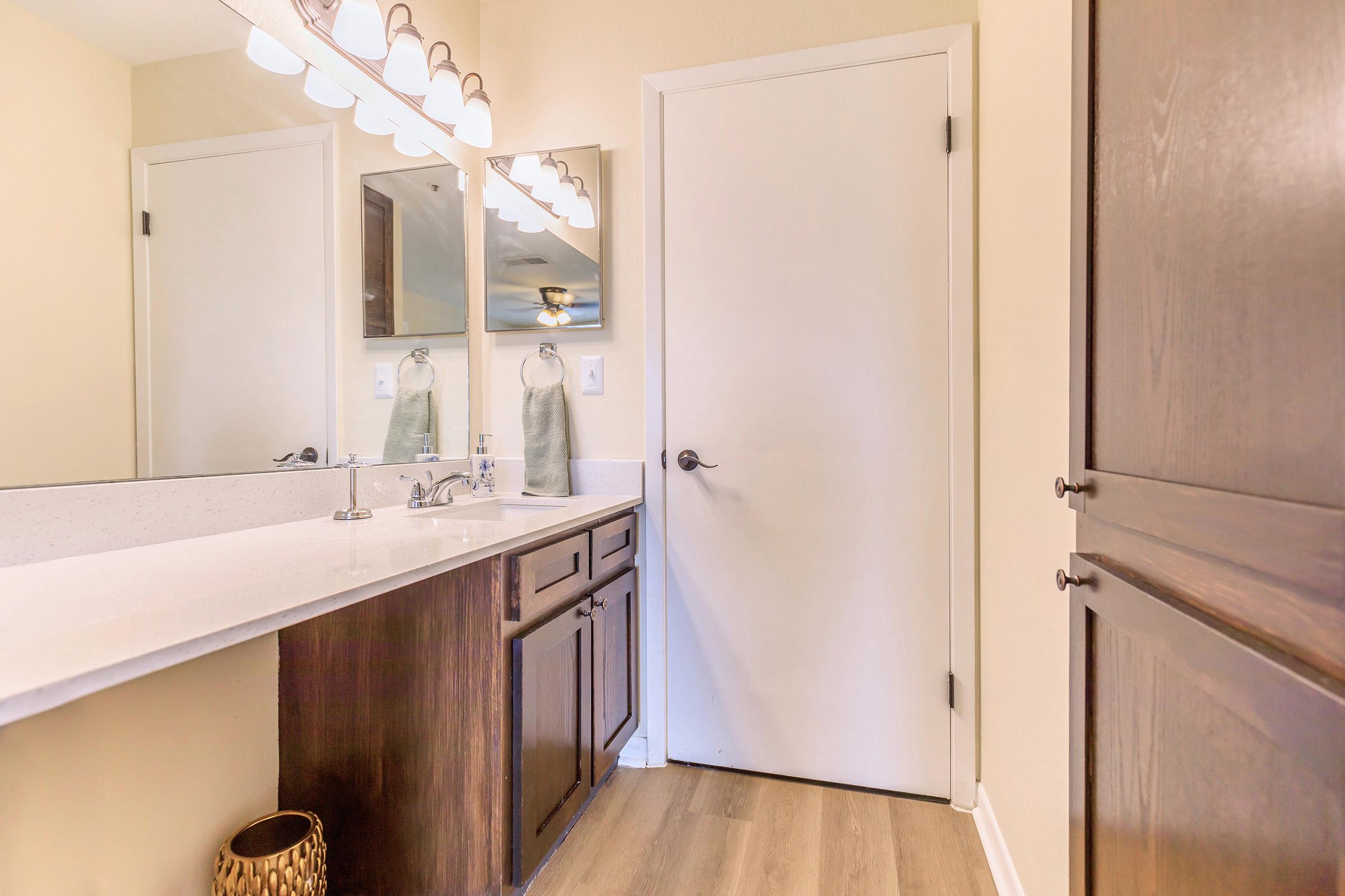 Brightly lit bathroom featuring a modern vanity with a white countertop, dual mirrors, and a faucet. A closed door is visible in the background, alongside dark wood cabinetry. The floor has a light wood appearance, and a woven basket is positioned near the vanity. The walls are painted a soft beige hue.