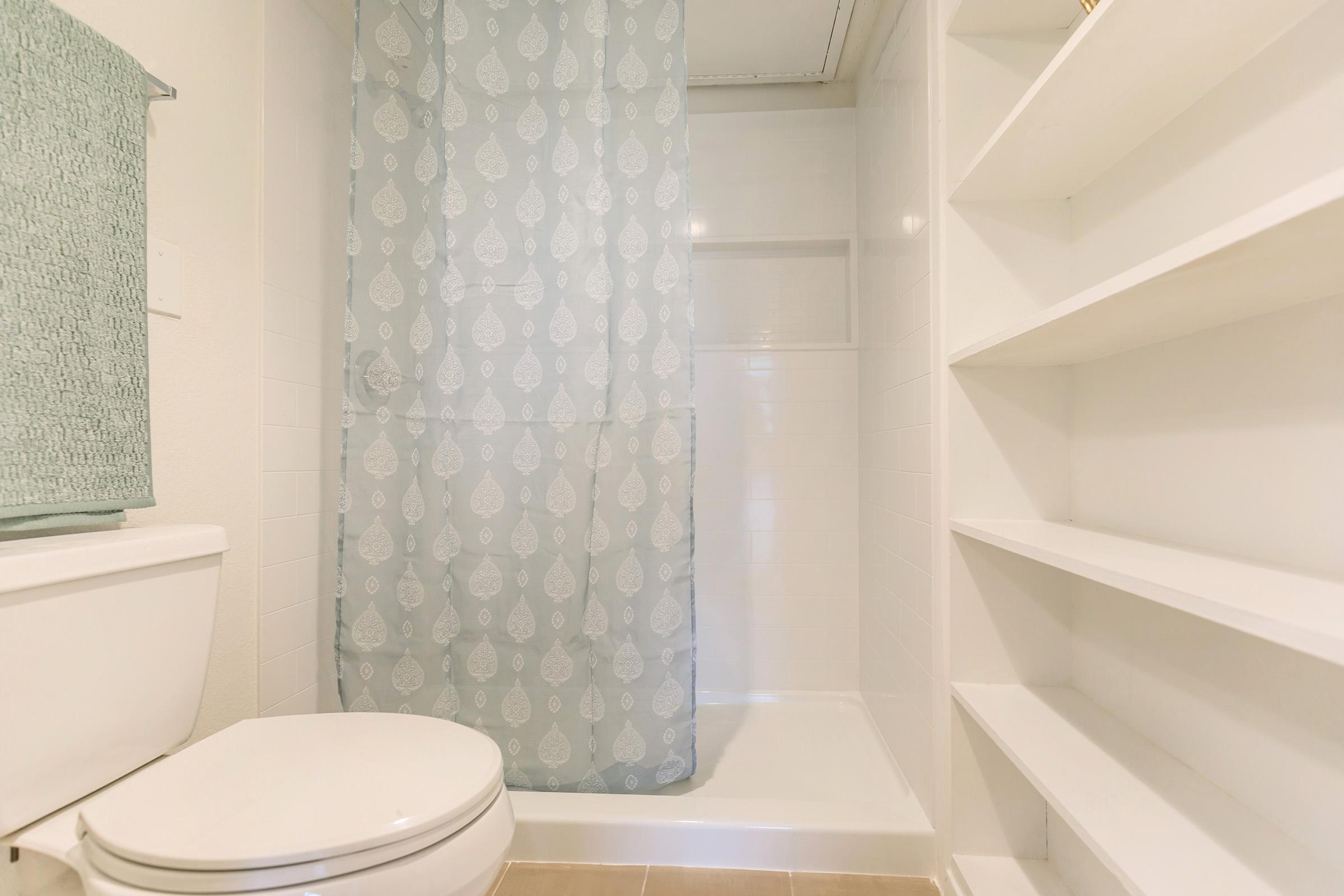 A clean bathroom featuring a white toilet, a shower with a light blue patterned curtain, and built-in white shelves. The walls are also white, and there is a soft green towel hanging nearby. The overall atmosphere is bright and minimalistic.