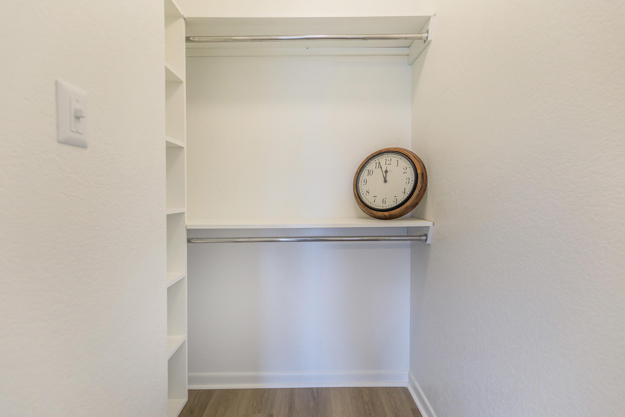 A minimalistic closet featuring two metal hanging rods and a wooden clock placed on a shelf. The walls are painted white, and the floor has a light wooden finish, creating a clean and spacious appearance. There are also built-in shelves on one side of the closet.