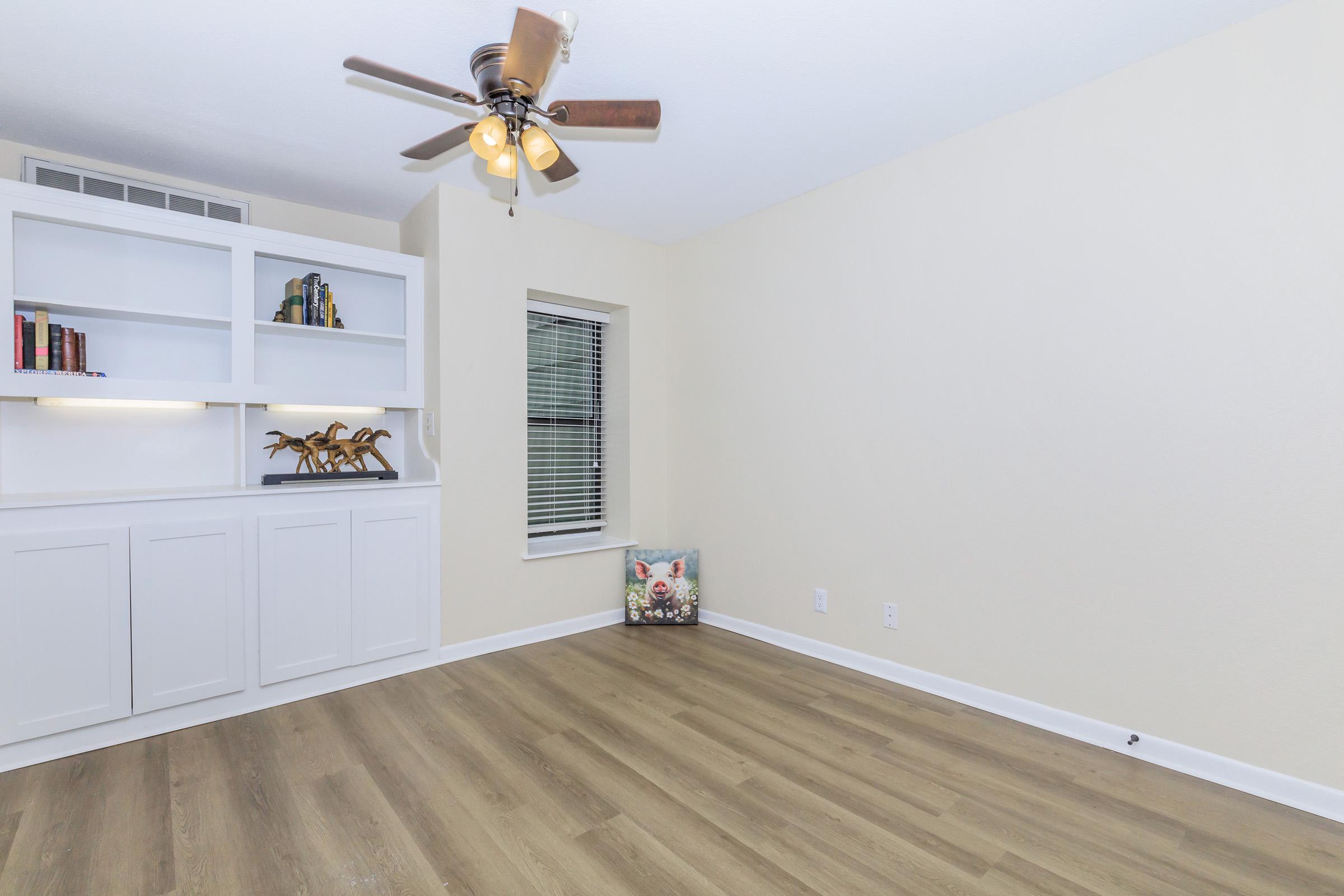 A spacious, empty room with light-colored walls and wooden flooring. There is a ceiling fan with light fixtures, a built-in bookshelf with a few books and decorative items, and a window with blinds. A decorative pillow is placed on the floor near the wall, contributing to the room's inviting atmosphere.