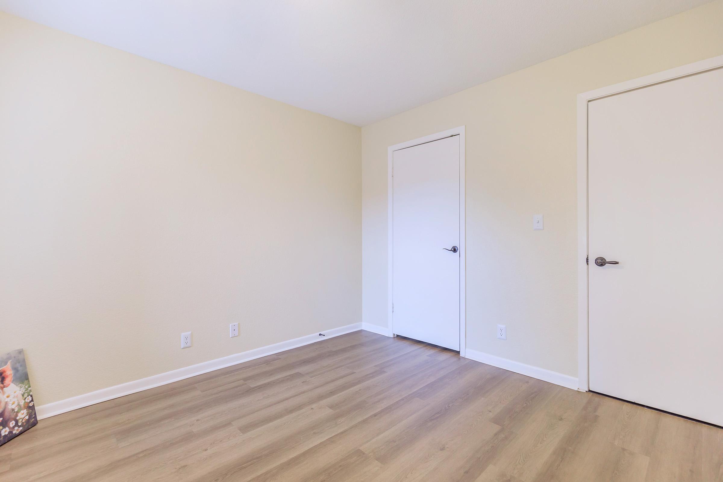 A minimalist room with light-colored walls and a wooden floor. There are two closed doors on the right side, and a small decorative item or painting partially visible on the left side. The overall atmosphere is clean and simple, suggesting a ready-to-use space.