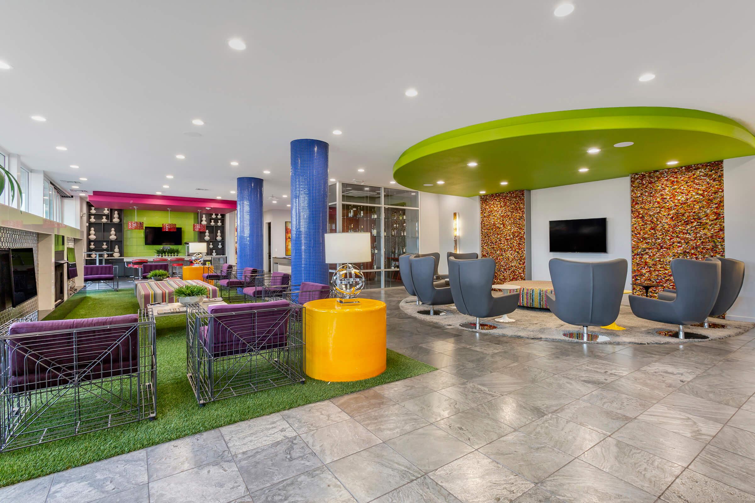 A modern and stylish lounge area featuring colorful furniture, including purple seating, bright yellow tables, and green accents. It has a green carpeted section, large windows providing natural light, and decorative wall features with vibrant patterns.