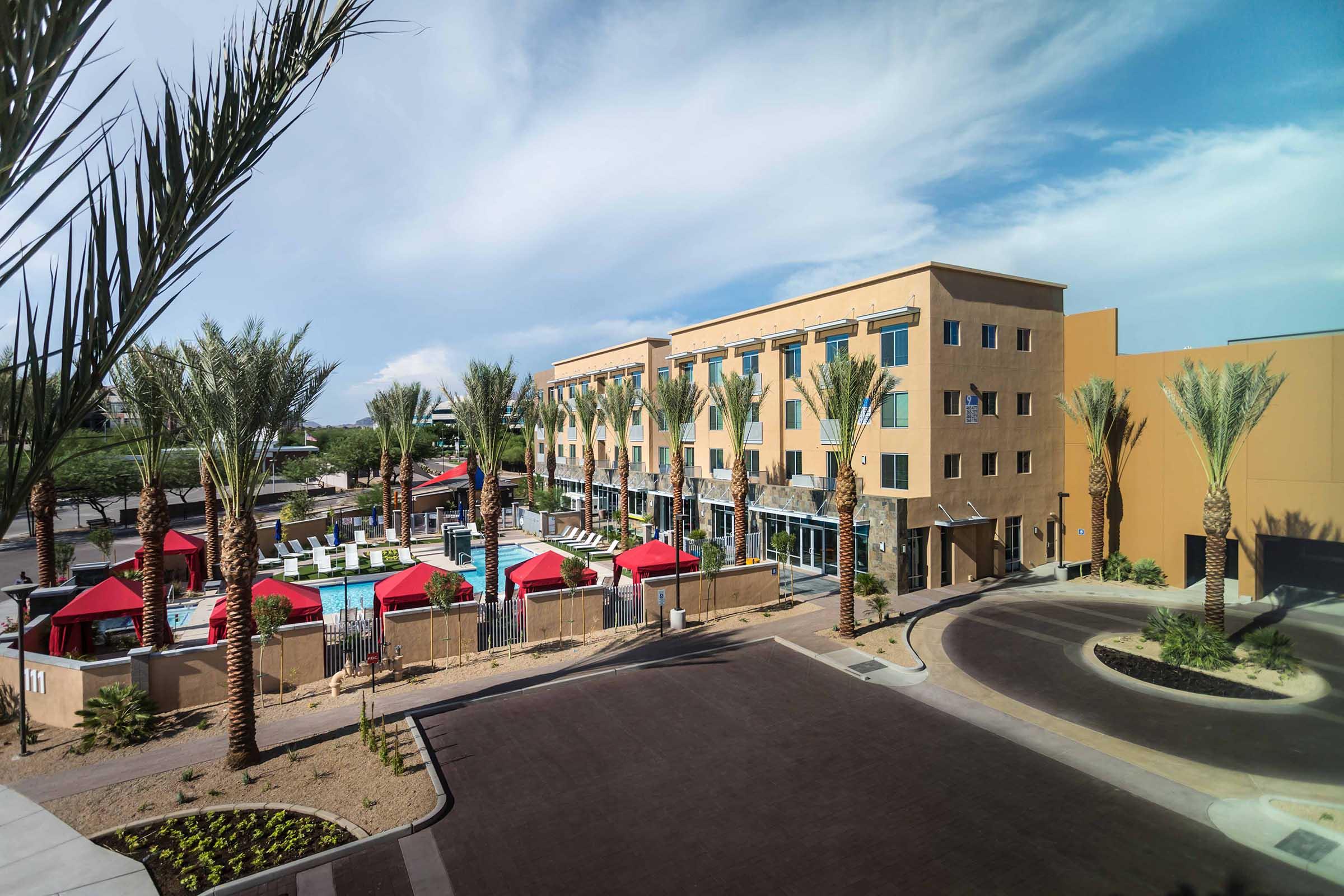 A modern hotel building with multiple floors, surrounded by palm trees and a pool area featuring red cabanas.