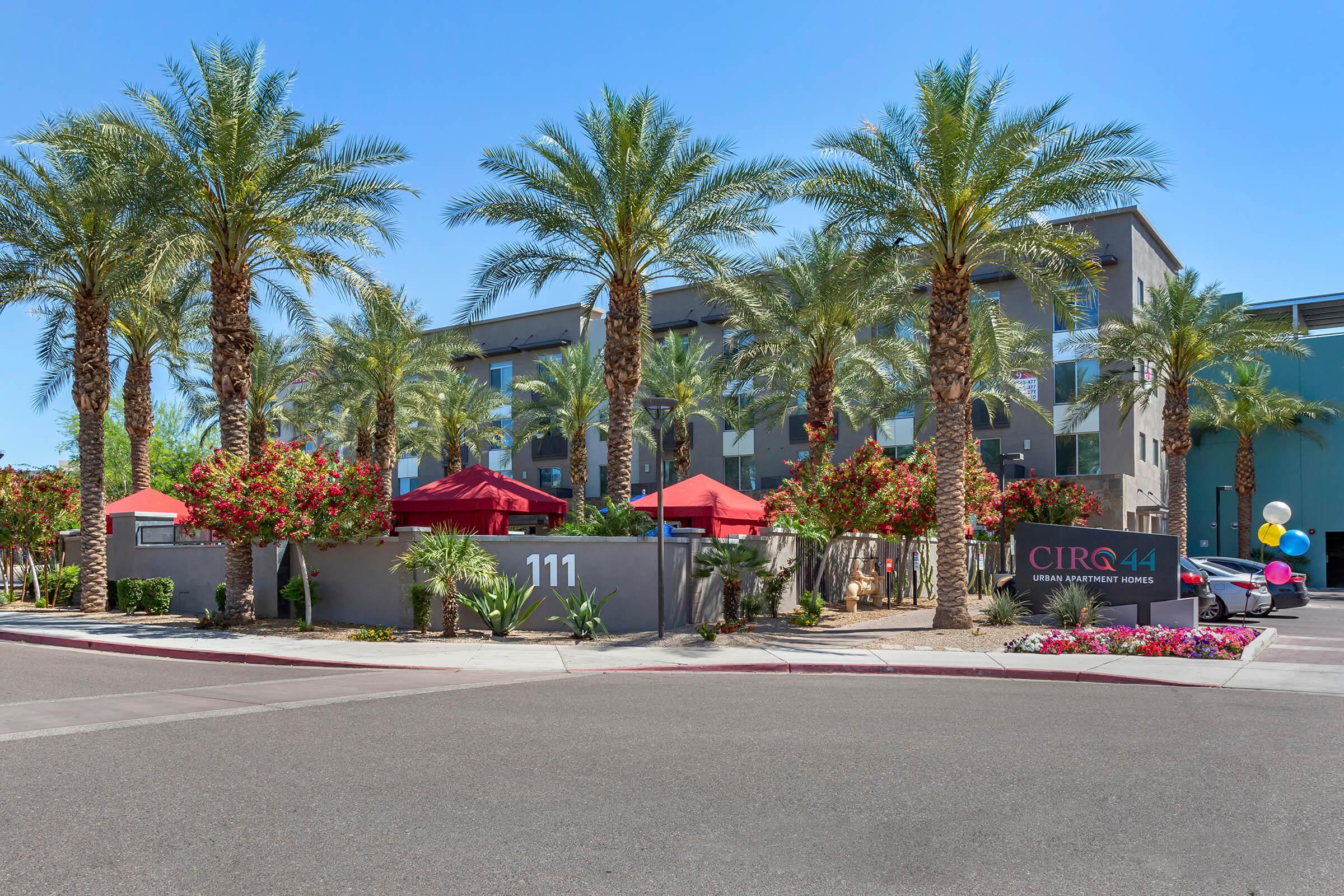 Modern apartment complex entrance with palm trees and colorful landscaping. Red awnings provide shade over outdoor seating areas.