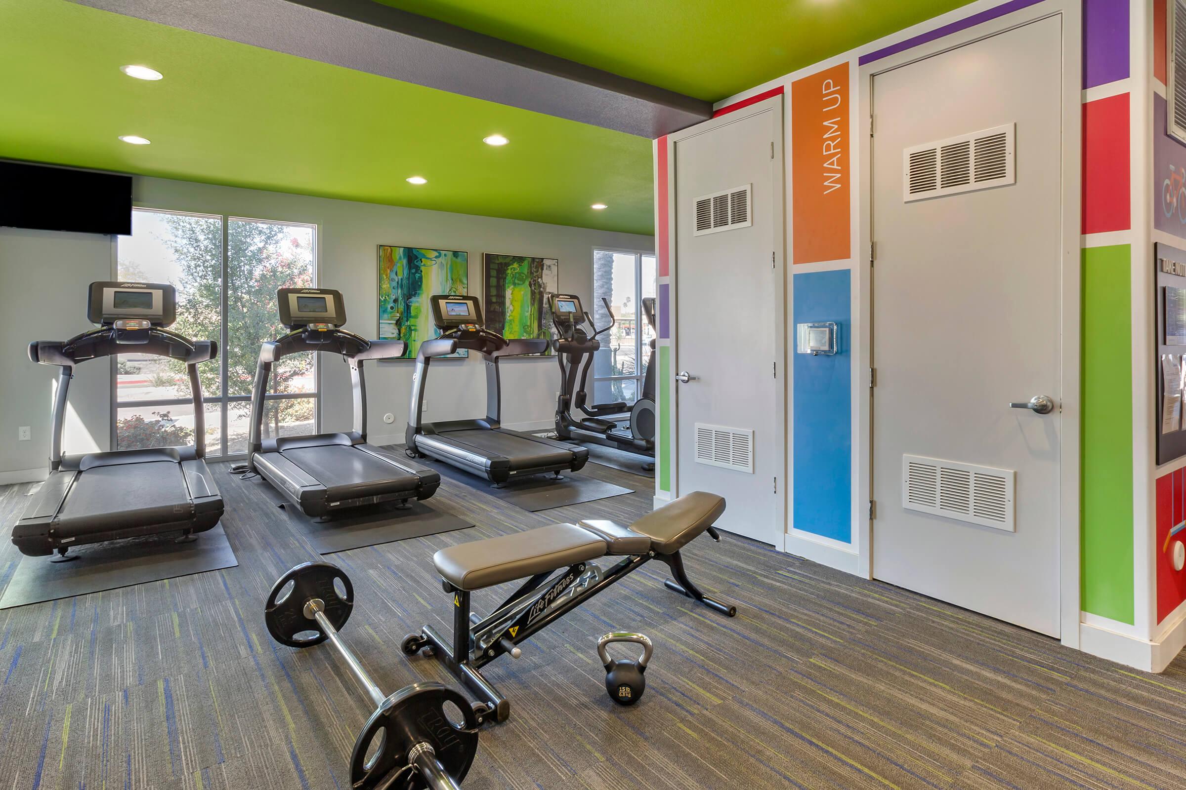 A modern gym space featuring three treadmills, a weight bench with a barbell, and colorful walls.