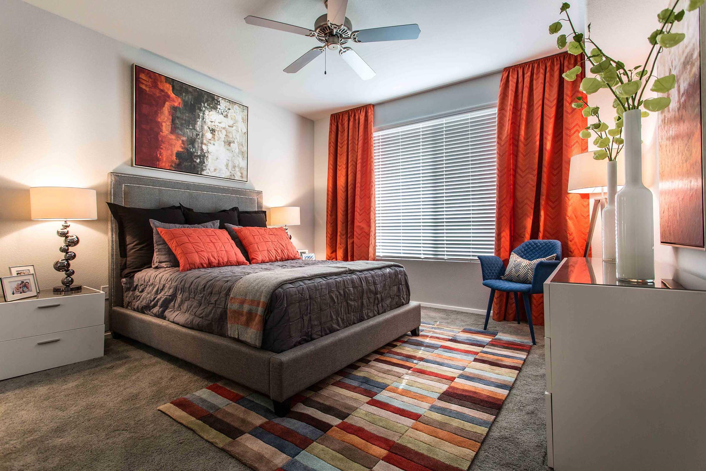 A well-decorated bedroom featuring a queen-sized bed with multiple decorative pillows, an abstract painting above the headboard, and orange curtains.