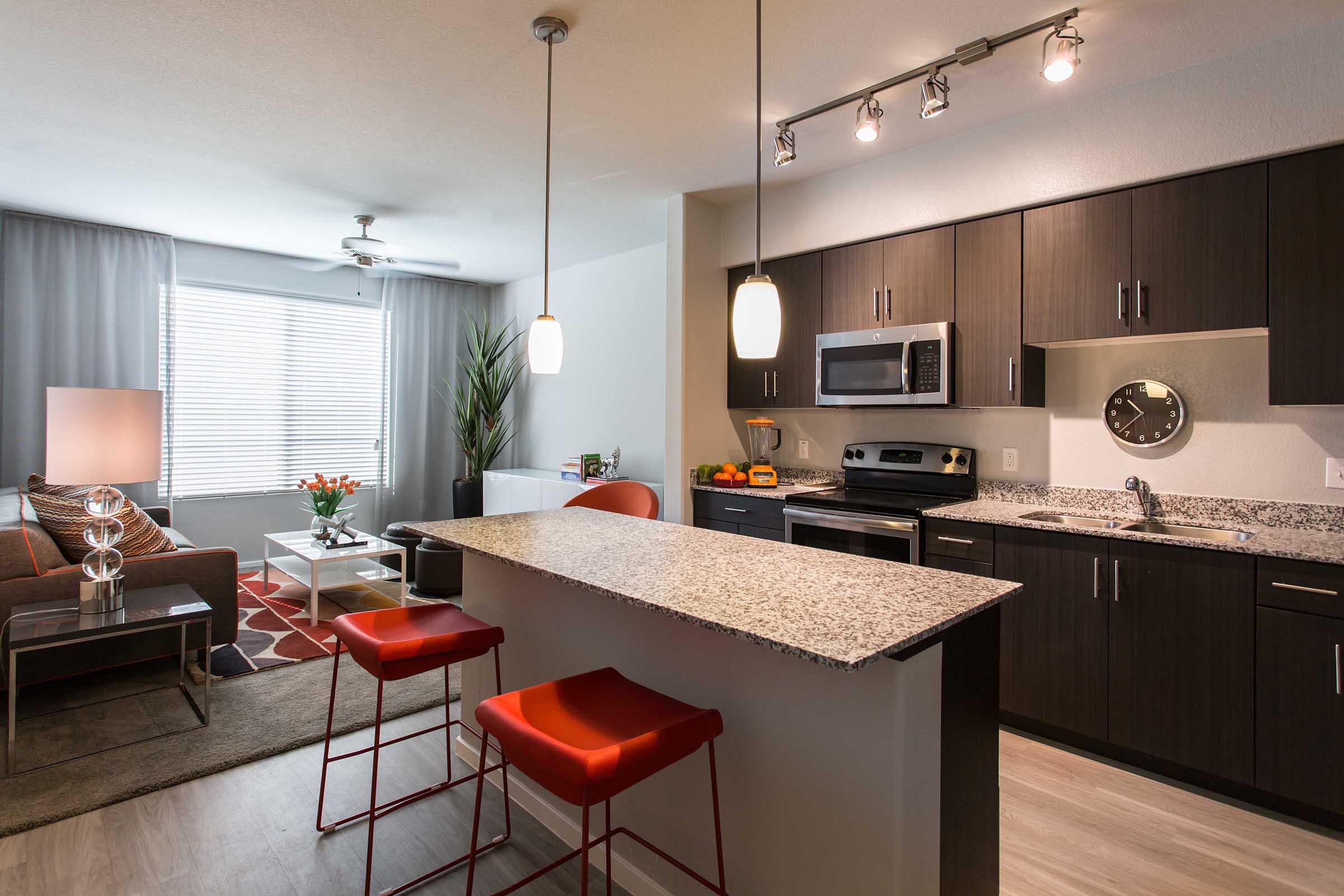 Modern kitchen and living area in an apartment. The kitchen features dark cabinetry, stainless steel appliances, and a granite island with red bar stools.