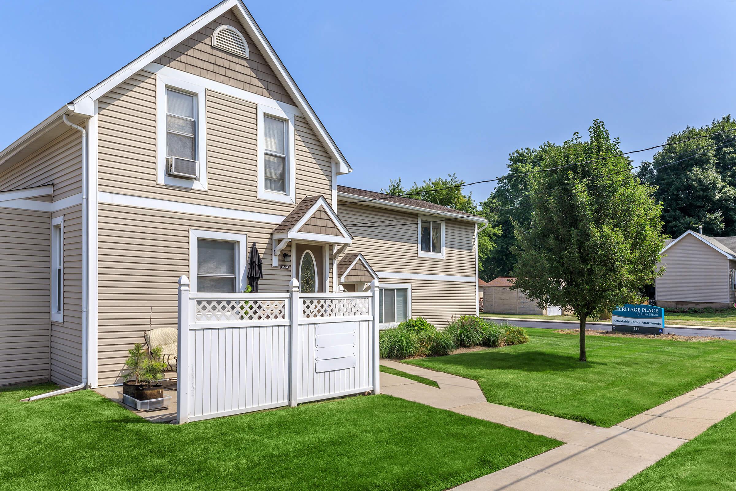 A two-story house with light beige siding, featuring white trim and a picket fence. The front yard is well-maintained with green grass and minimal landscaping. A pathway leads to the front door, and there's a small tree near the fence. The street is visible in the background with a sign for "Shady Place."