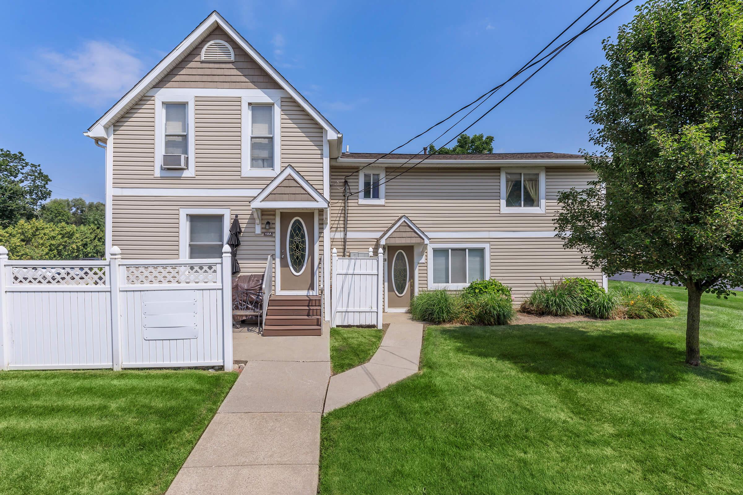 A two-story house with beige siding, featuring a white picket fence, a concrete walkway leading to the front door, and well-maintained green lawns on either side. There are windows with decorative trim and greenery around the property, under a clear blue sky.