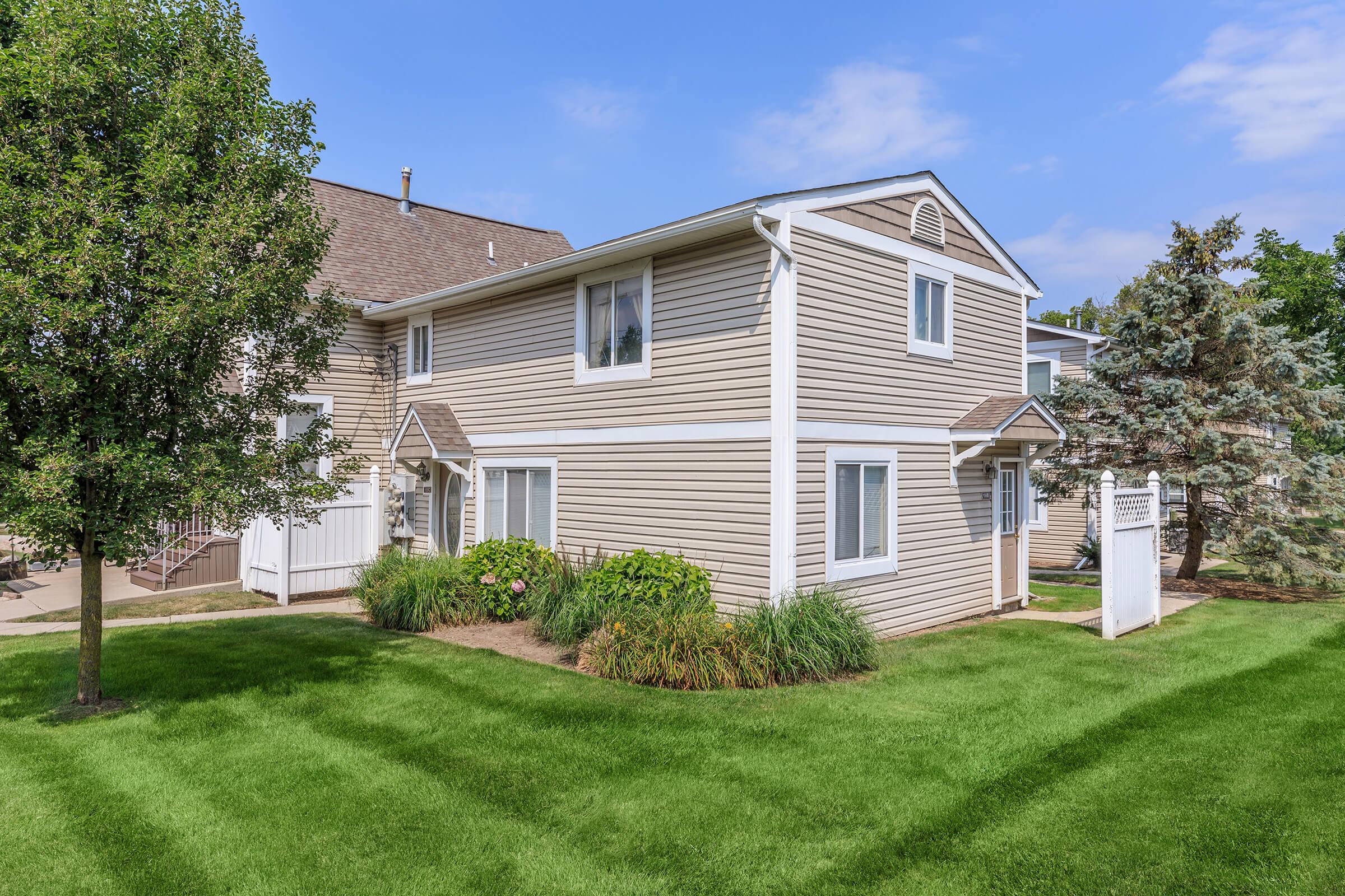 A two-story residential building with beige siding surrounded by green grass and shrubs. There are large windows on each floor, and a sign is positioned near the entrance. Trees provide some shade, and the sky is clear with a few clouds.