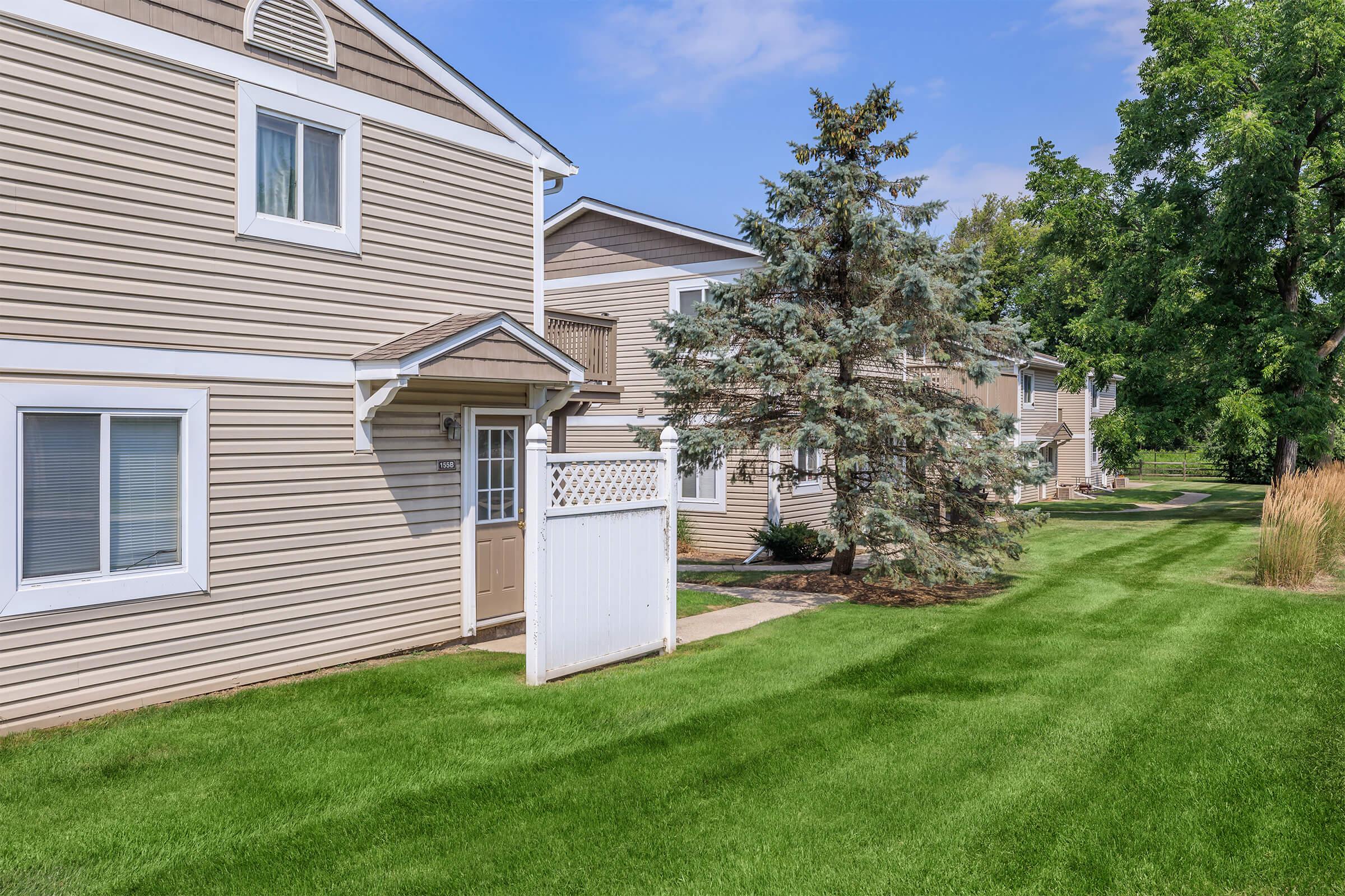 Well-maintained residential area featuring two-story beige homes with white trim. Neatly trimmed green grass and a small tree are visible, along with a white fence in the foreground. The scene is bright and sunny, showcasing a peaceful neighborhood atmosphere.