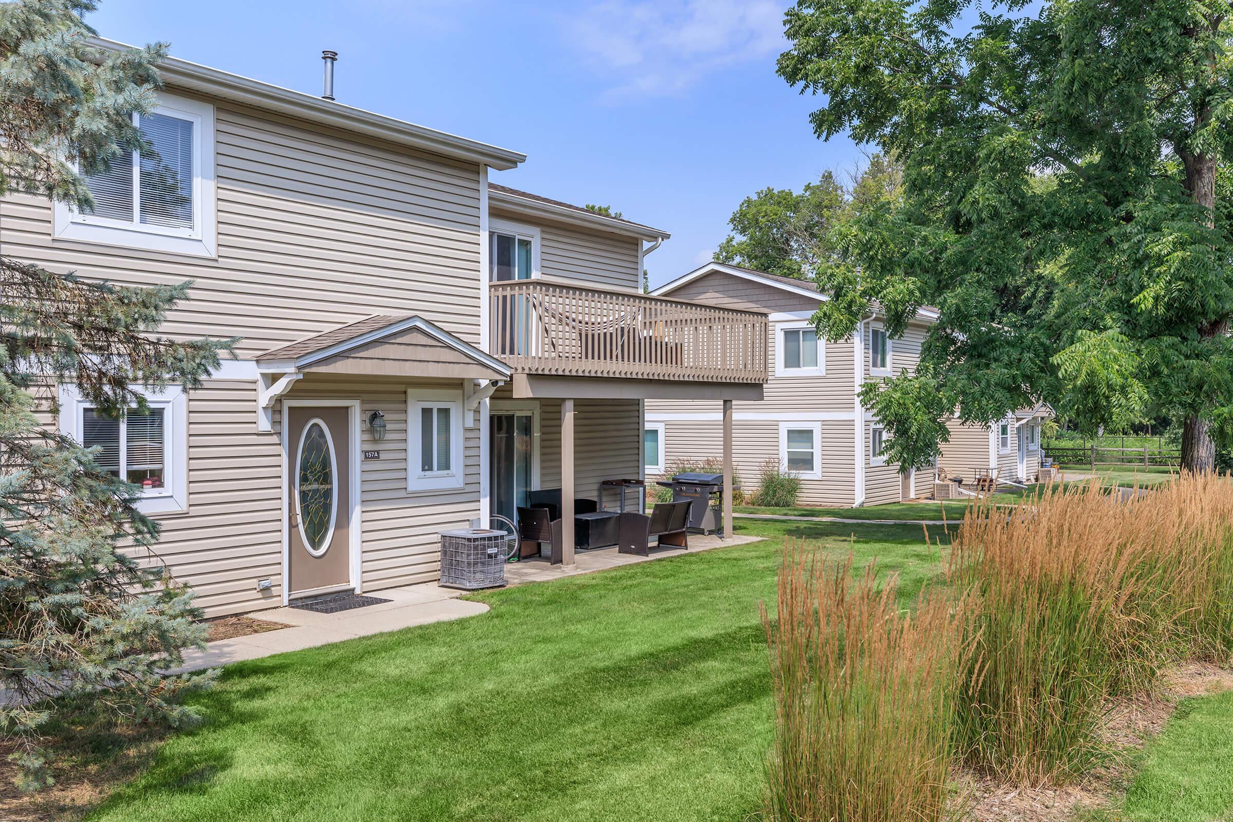 A view of a two-story house with beige siding, featuring a balcony and a porch area with outdoor seating. The surrounding area includes well-maintained green grass and tall ornamental grasses, with trees providing shade in the background under a blue sky.