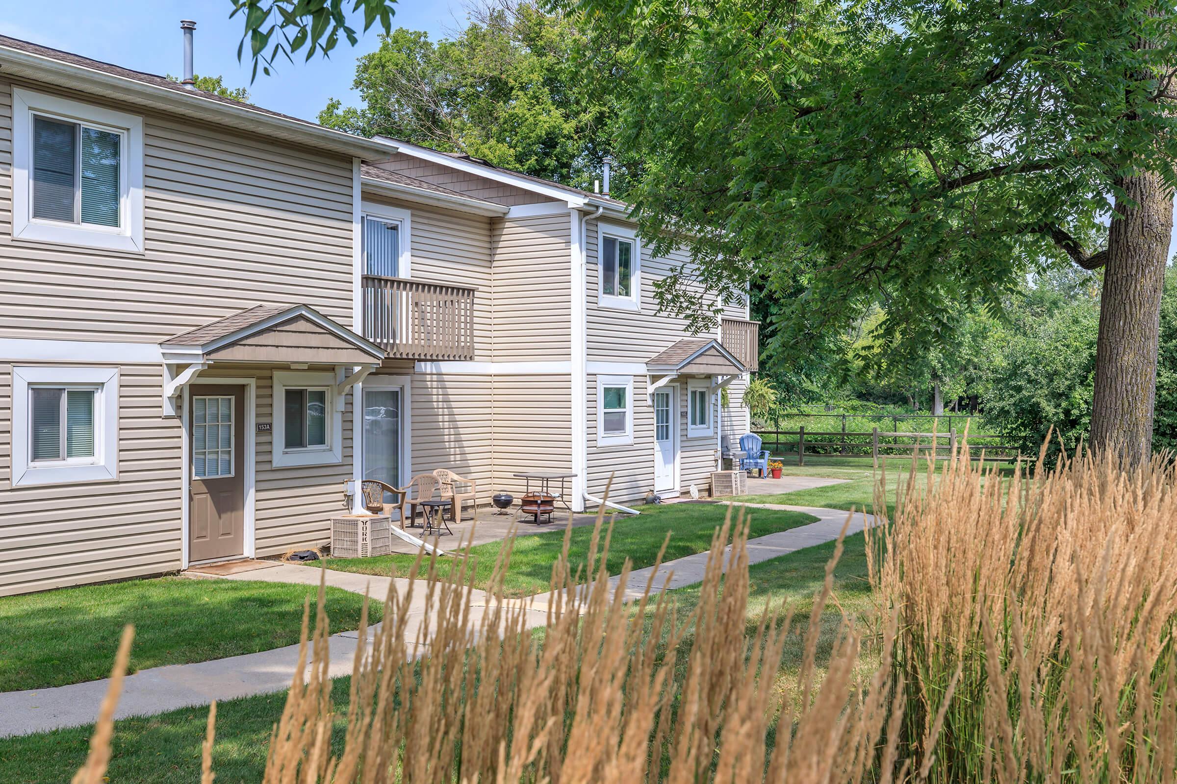 A view of a multi-unit residential building with light-colored siding, featuring balconies and a pathway alongside a grassy area. In the foreground, tall grasses are visible, and lush trees provide shade around the property, creating a serene outdoor setting.