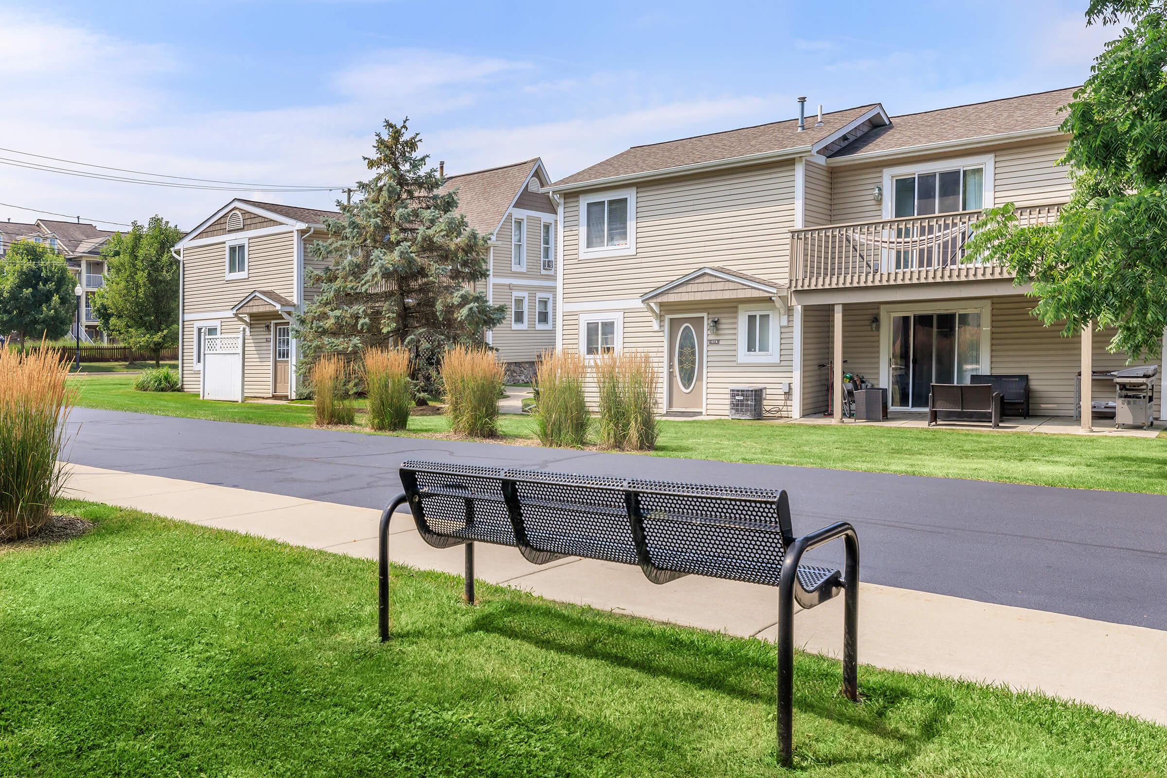 A scenic view of a residential neighborhood featuring two two-story houses with balconies, surrounded by green lawns and ornamental grasses. A paved pathway leads to a metal bench, providing a spot to sit and enjoy the outdoors. The sky is clear with a few clouds, reflecting a pleasant day.