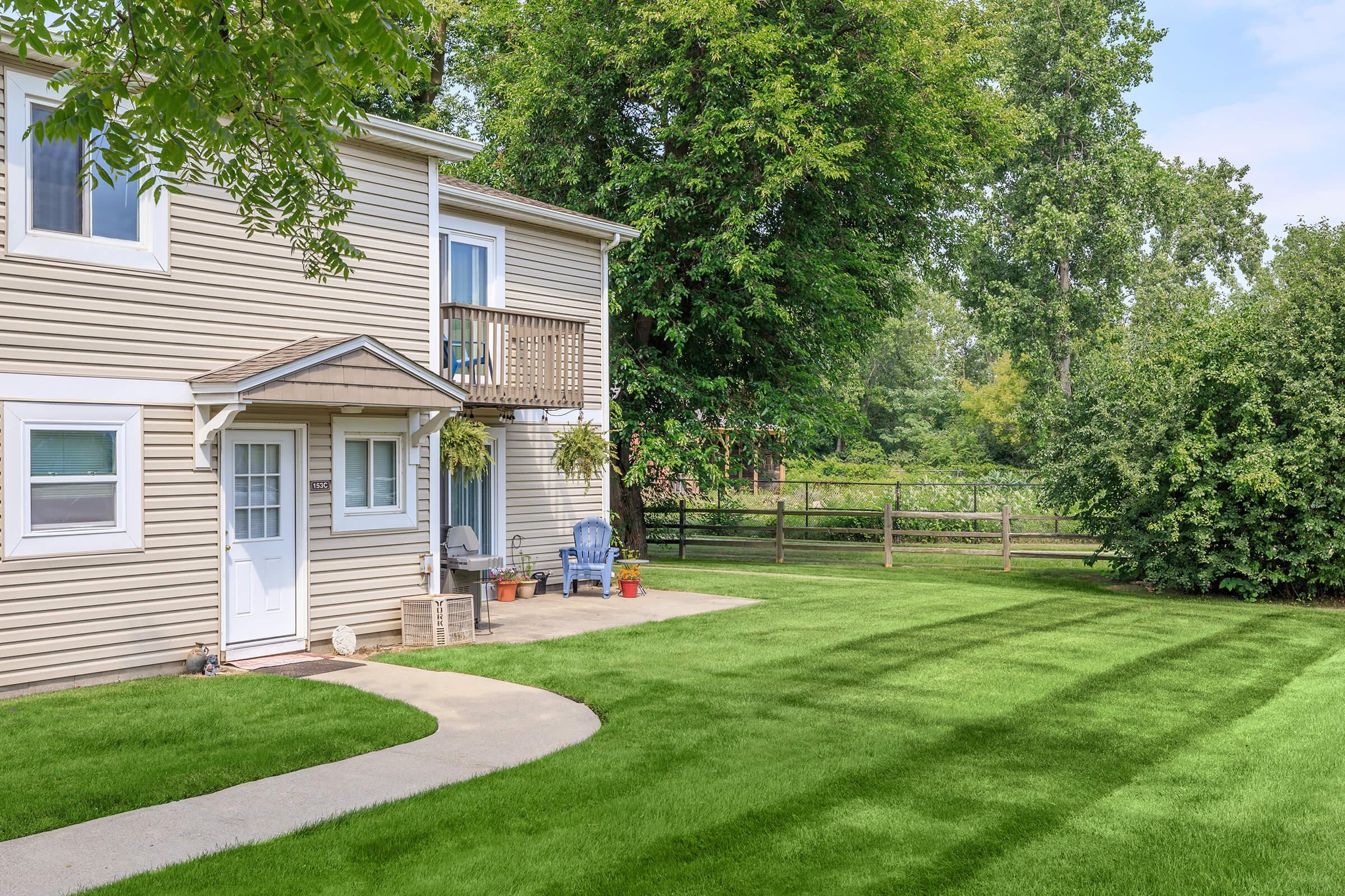 A cozy two-story home with a beige exterior and a small balcony, surrounded by lush green grass and trees. Two blue chairs are positioned on the patio, and a pathway leads to the entrance. A wooden fence and additional greenery are visible in the background, creating a serene outdoor space.