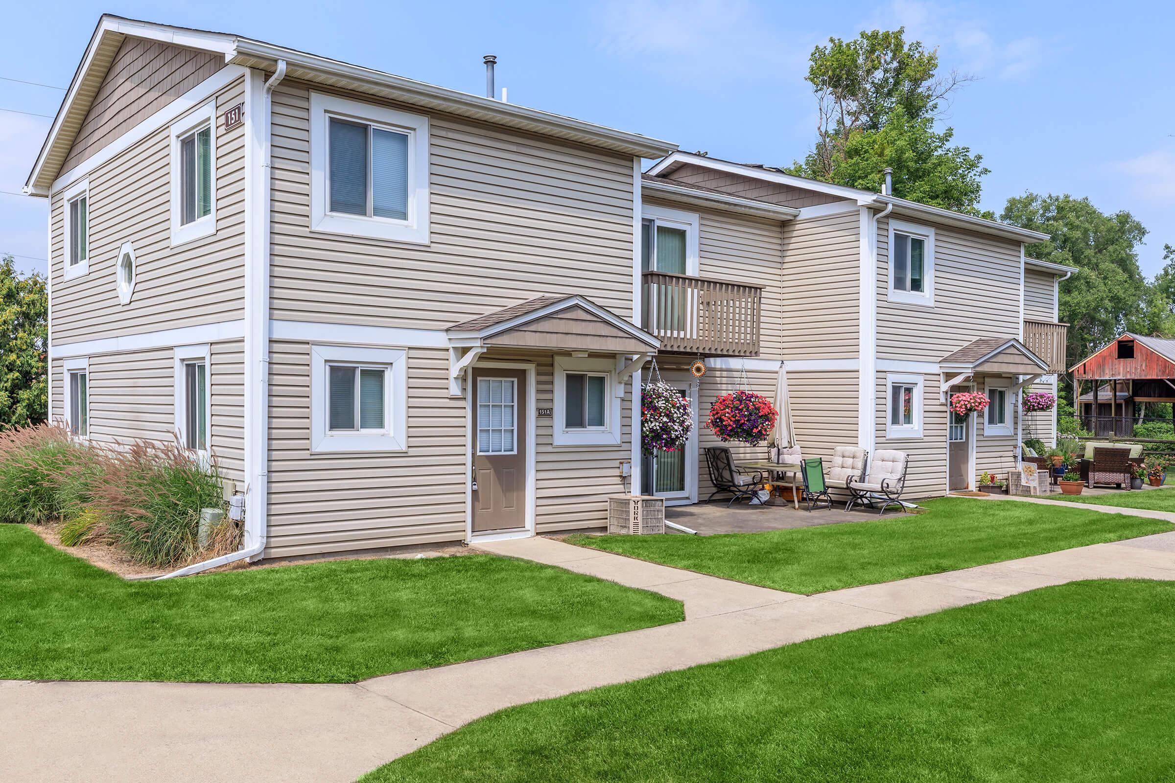 A two-story beige apartment building with multiple windows, balconies, and flower pots. A well-maintained green lawn and a paved walkway lead to the entrance. There are outdoor sitting areas with chairs. In the background, a red barn is partially visible among trees.