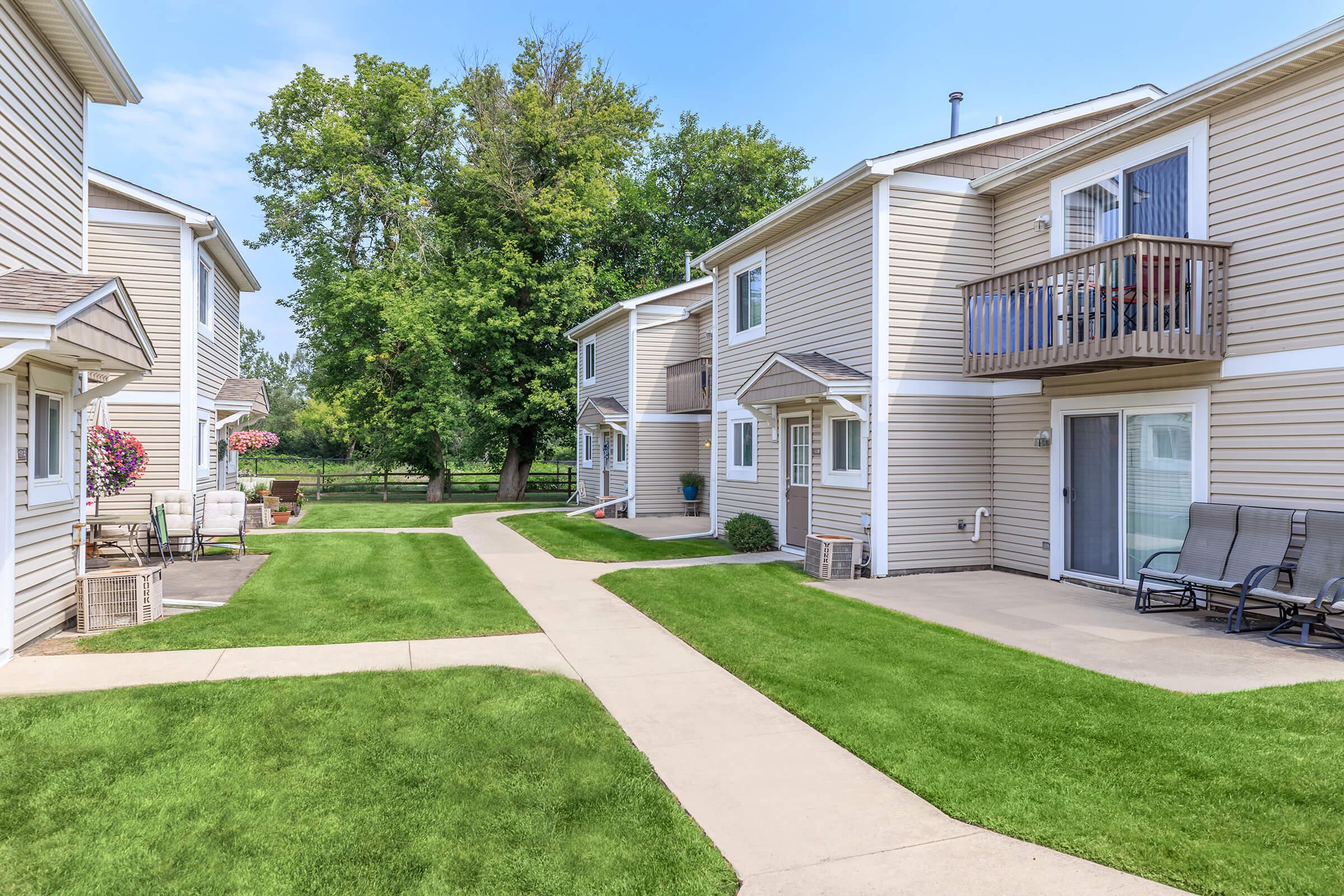 A peaceful residential area featuring two-story apartment buildings with beige siding. Neatly manicured lawns with green grass and a concrete pathway connect the units. Flower pots adorn the outdoor spaces, and trees provide a natural backdrop, creating a serene neighborhood atmosphere.