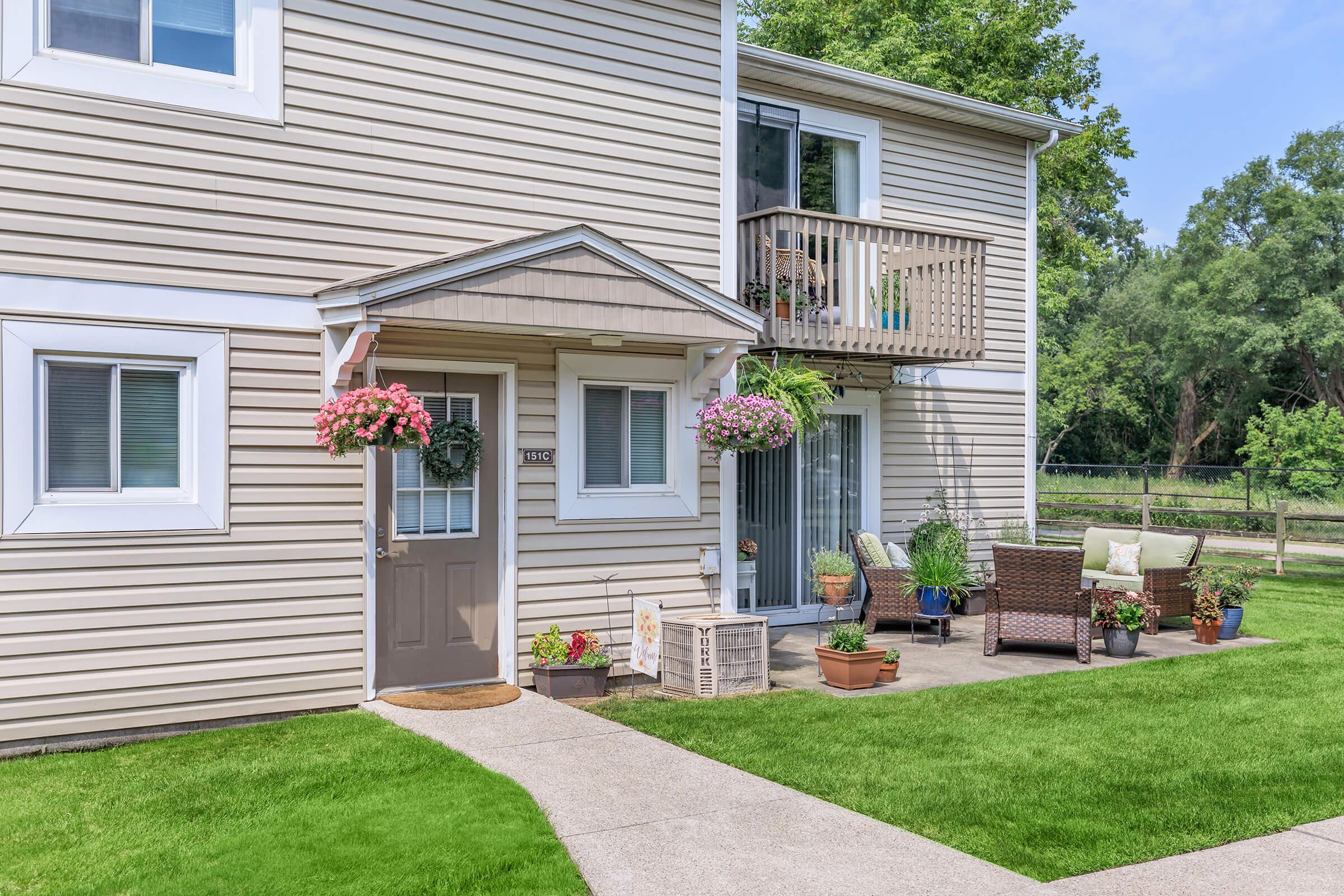 A well-maintained two-story apartment building with beige siding. The entrance features a small porch adorned with hanging flower baskets. There are potted plants and outdoor furniture on the patio, surrounded by green grass and trees in the background, creating a welcoming atmosphere.