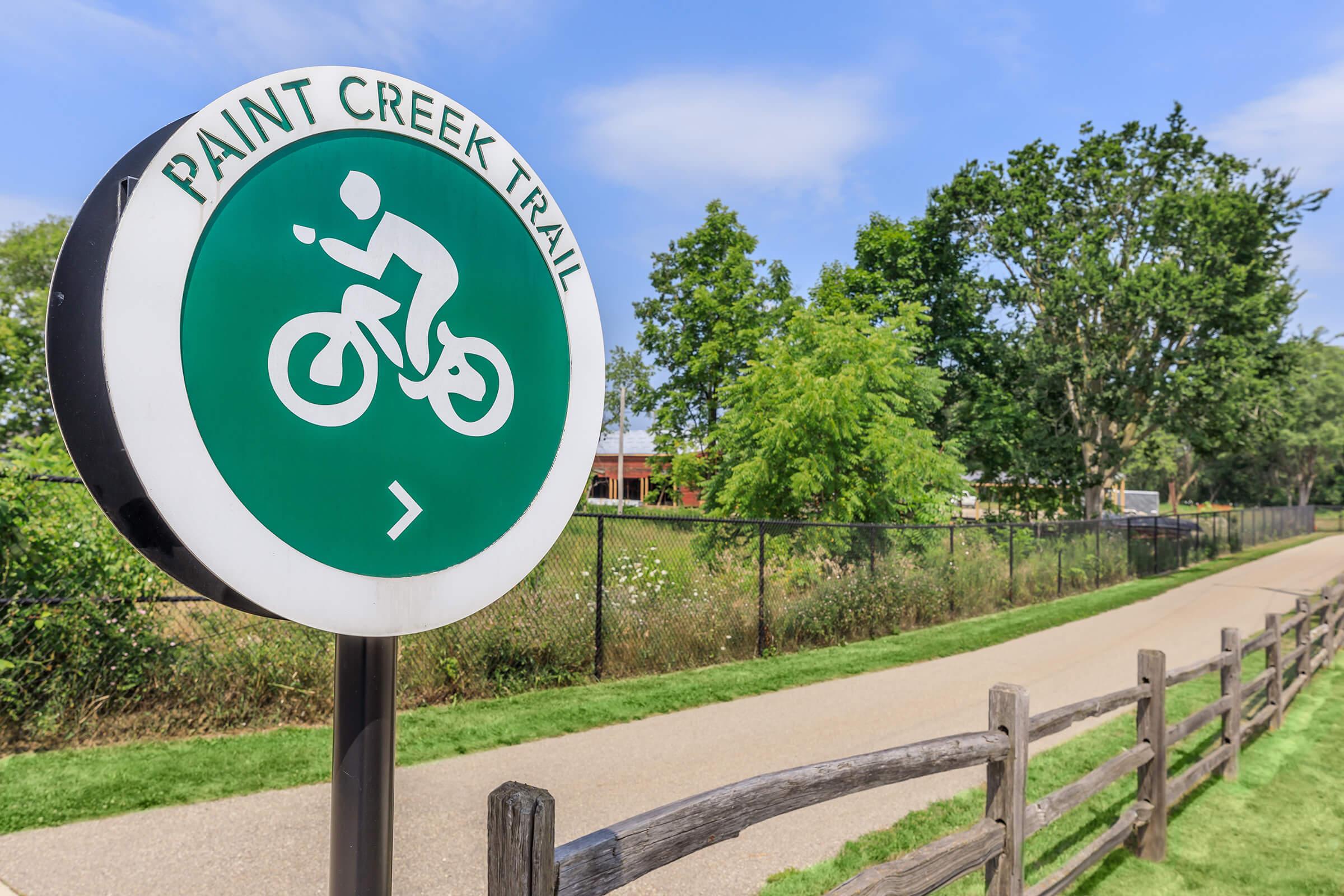 Sign for Paint Creek Trail featuring a bicycle icon, with a pathway and trees in the background. A wooden fence lines the trail. The sky is clear and blue, indicating a pleasant day for outdoor activities.