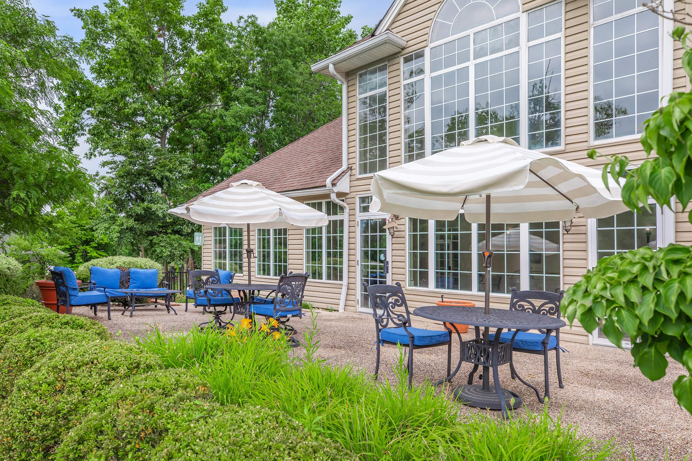 A spacious outdoor patio with blue cushioned chairs and brown tables under white umbrellas, surrounded by lush greenery. In the background, large windows of a house reflect the trees and sky, creating a serene and inviting outdoor seating area.