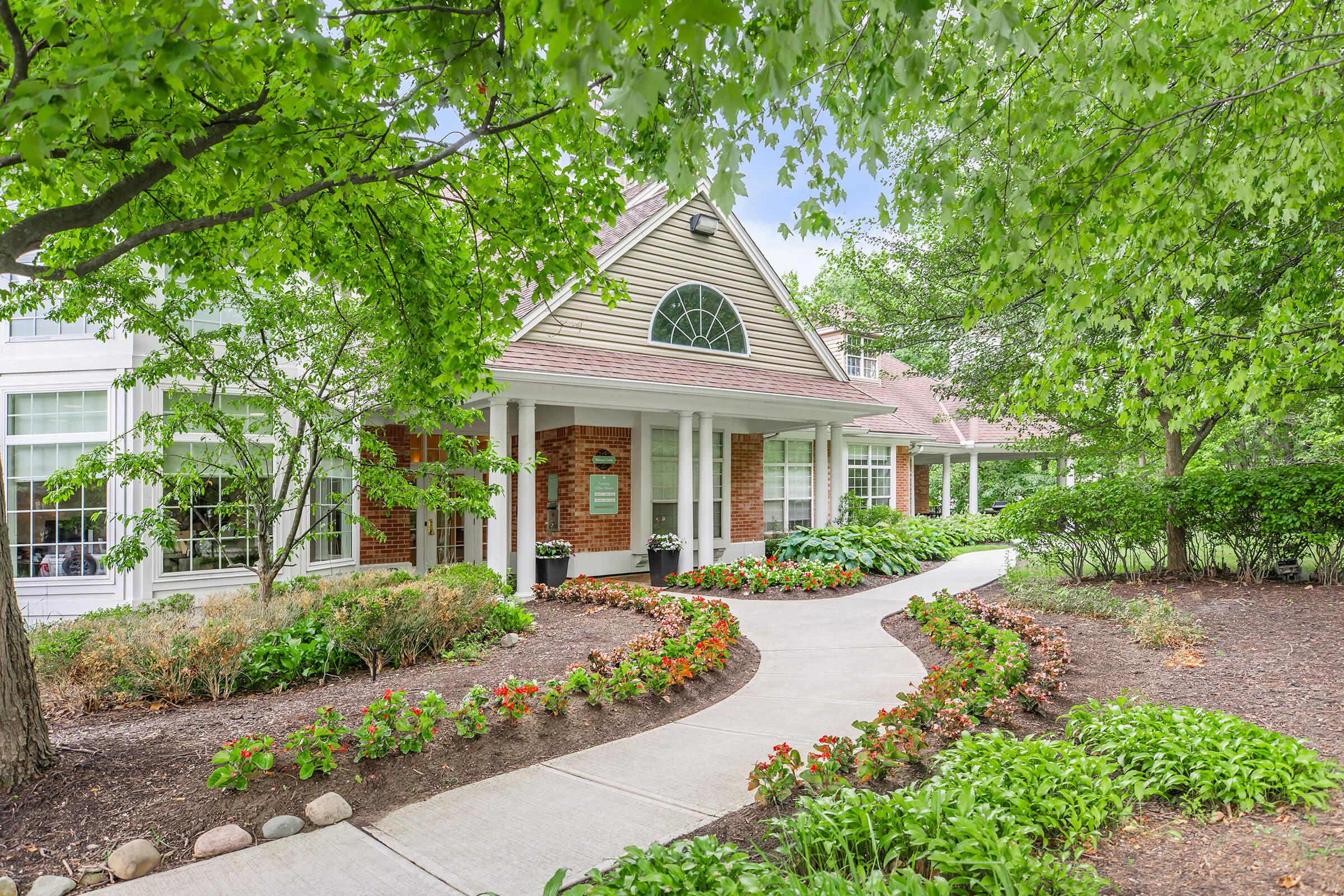 A charming building with a sloped roof surrounded by lush greenery. The pathway is lined with colorful flower beds, leading to the entrance, which features large windows and a welcoming porch. Trees overhead provide shade, creating a serene and inviting atmosphere.