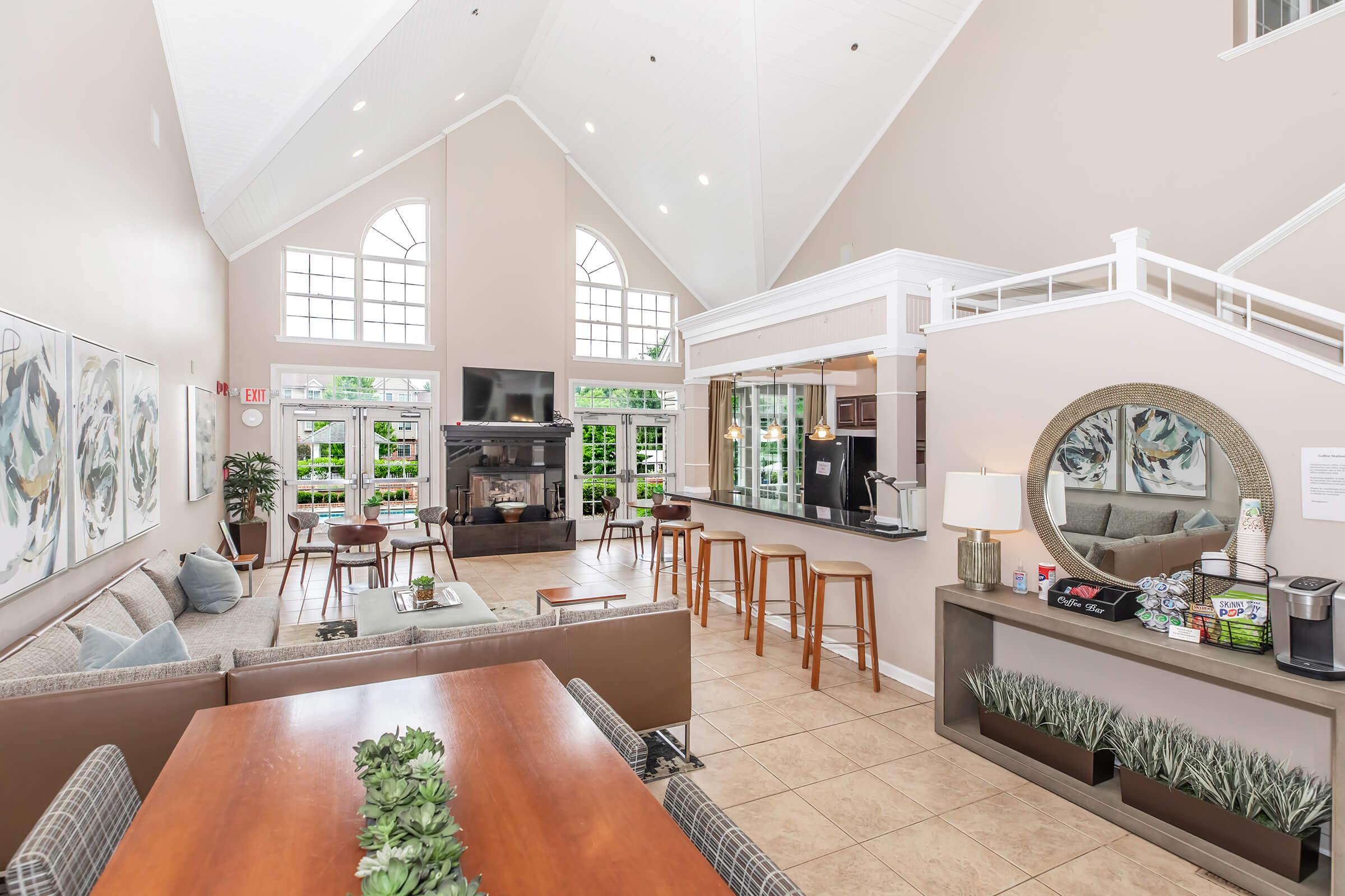 A spacious and modern common area featuring a high ceiling, large windows allowing natural light, comfortable seating with couches and chairs, a wood dining table, a bar with stools, and decorative plants. A cozy fireplace is visible, along with artwork on the walls and a coffee station.