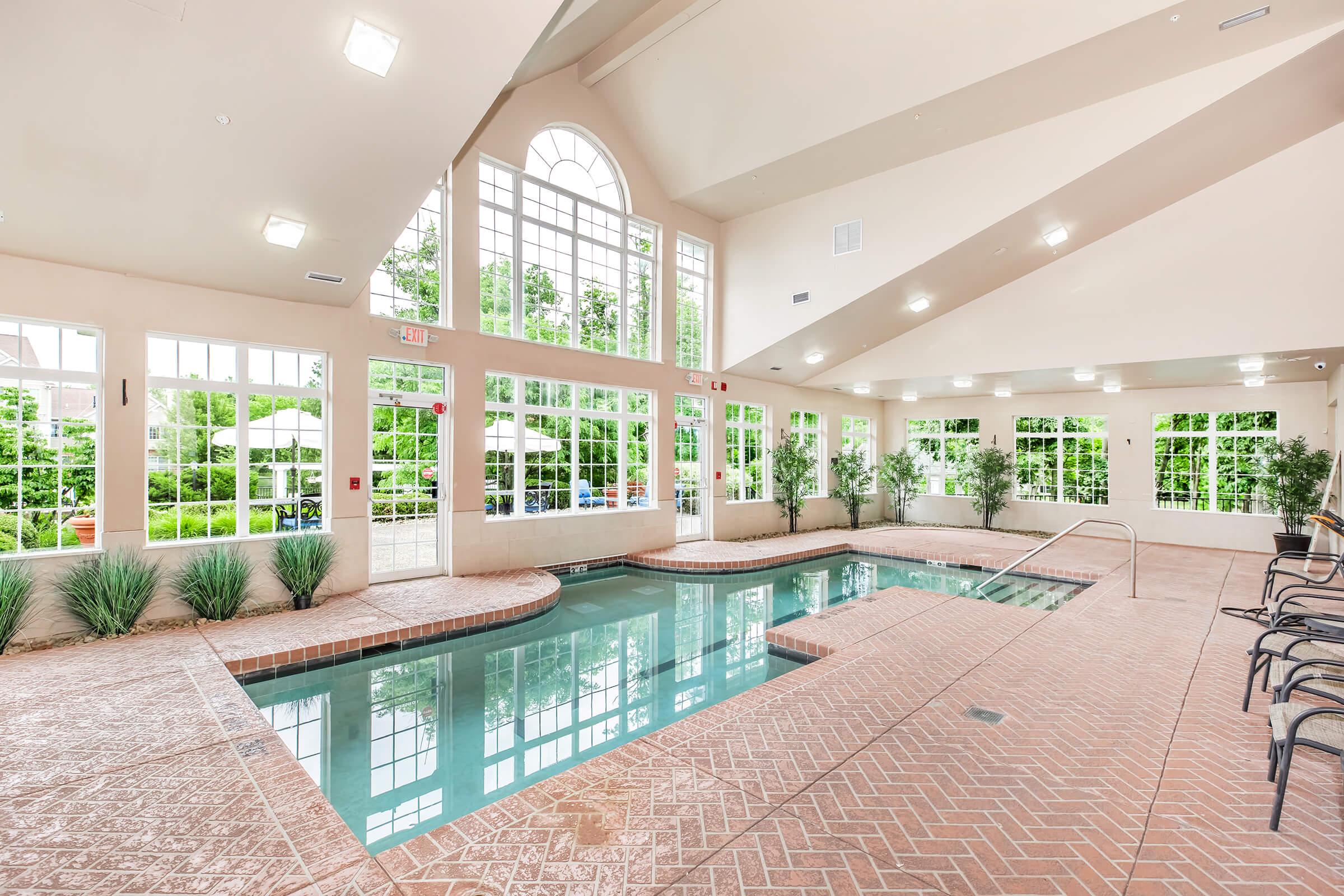 Indoor swimming pool area featuring large windows providing natural light, with a tiled pool, surrounding greenery visible outside, and several potted plants. Comfortable seating is arranged along the poolside.