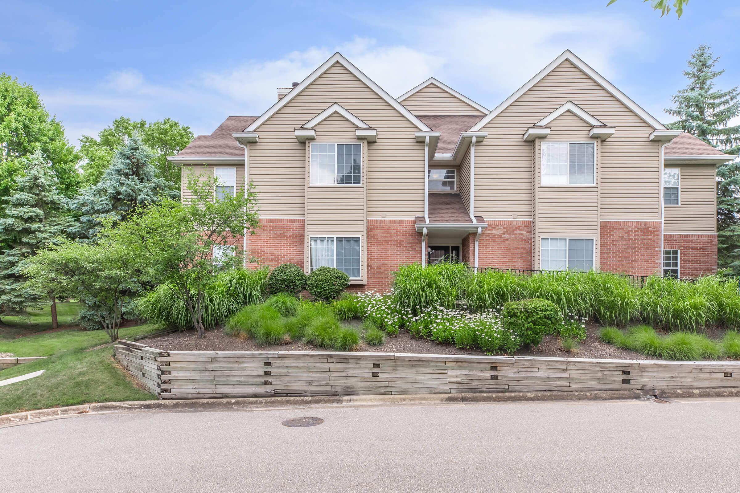Exterior view of a multi-unit residential building featuring a mix of beige and red brick siding. The building is set in a landscaped area with green bushes, small trees, and blooming flowers, surrounded by a gently sloped lawn. The sky above is clear with a few clouds, enhancing the inviting atmosphere.