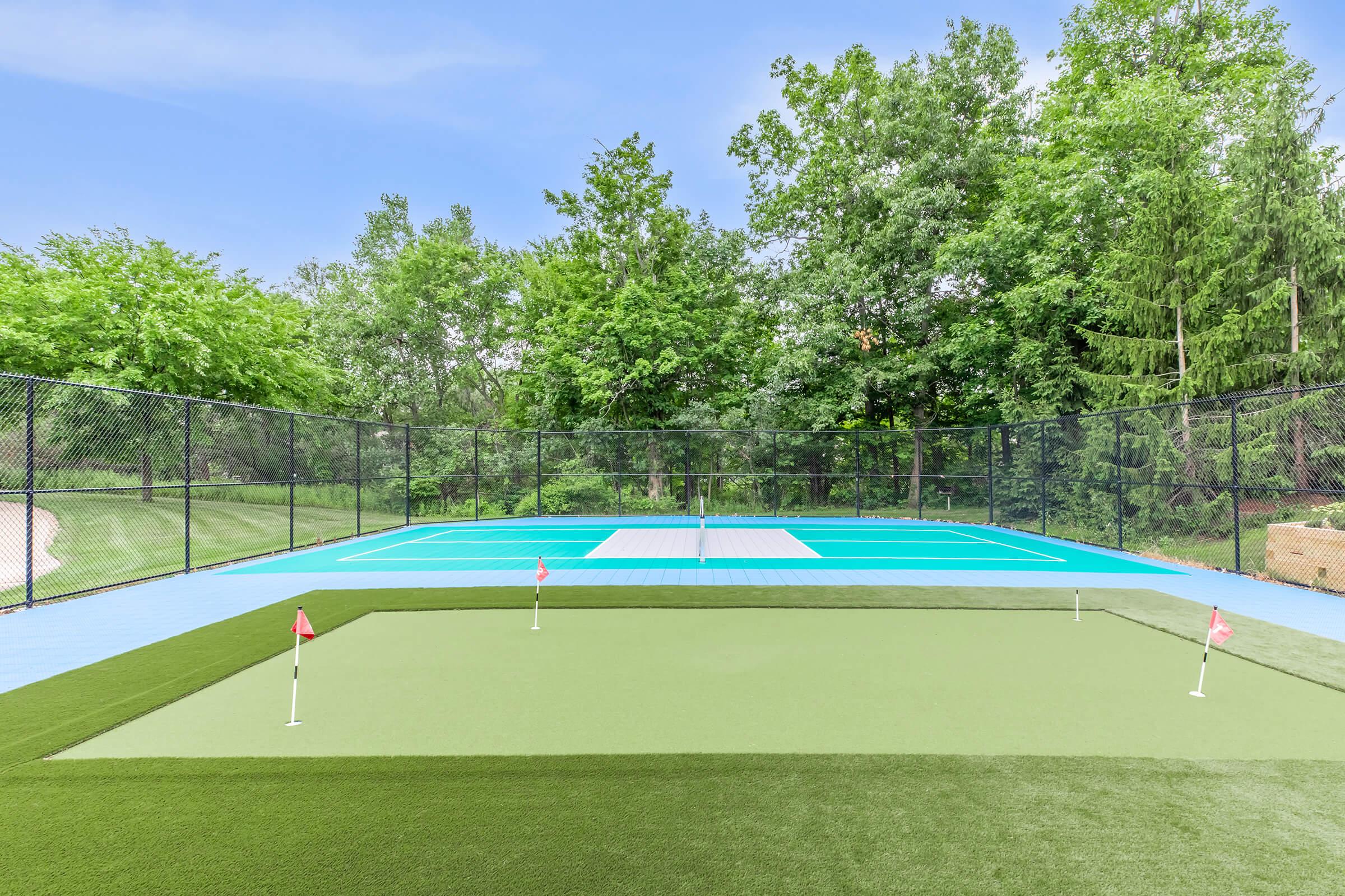 A bright outdoor sports area featuring two tennis courts surrounded by greenery. One court is painted blue and the other has a net, while a putting green with two holes and flags is in the foreground. The area is enclosed with a black mesh fence, creating a serene environment for playing sports.
