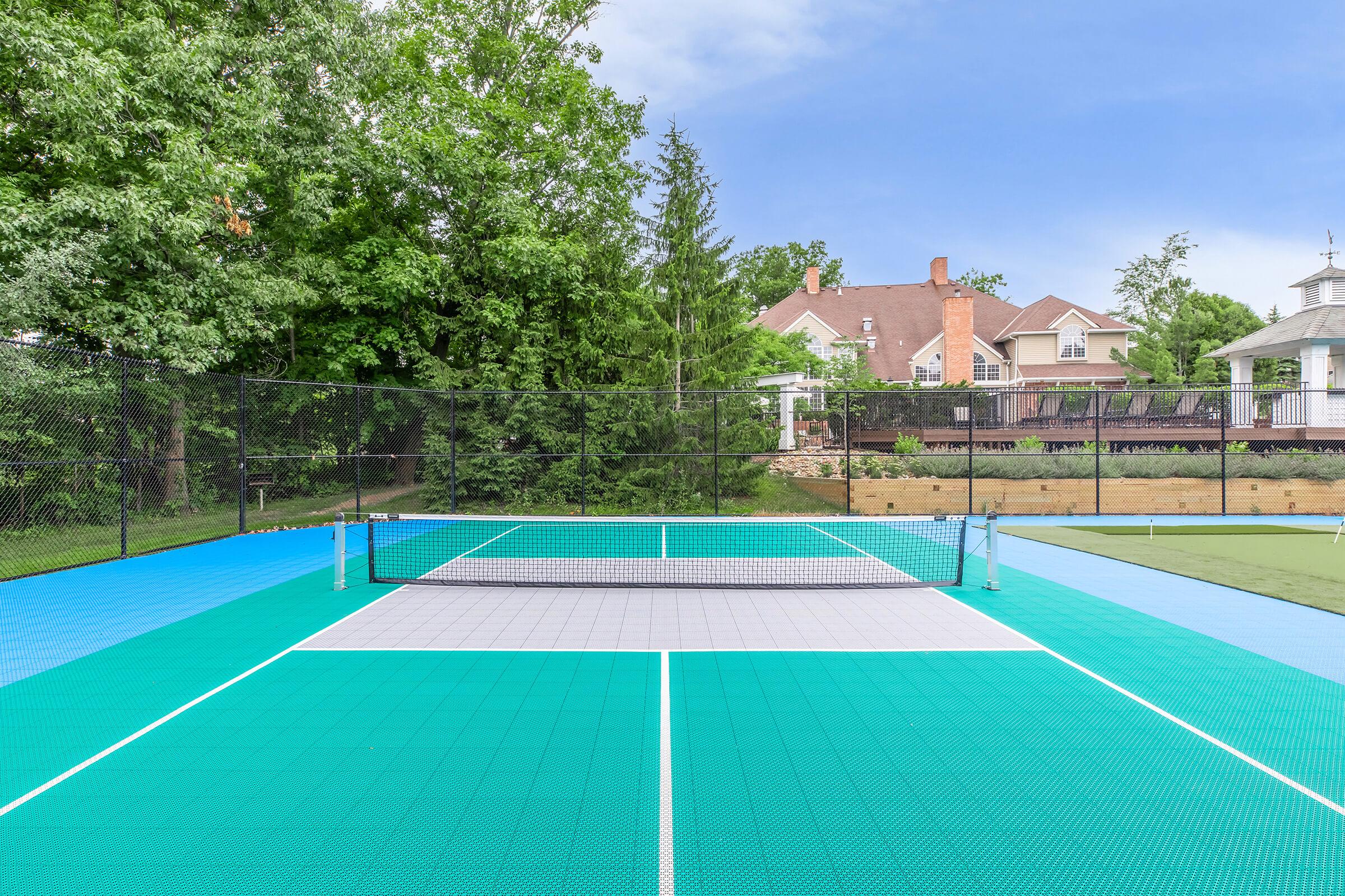 A view of a brightly colored tennis court featuring a green and blue surface. The net is set up in the middle, with surrounding trees and a residential area visible in the background. The scene is clear and sunny, providing a tranquil setting for a game of tennis.