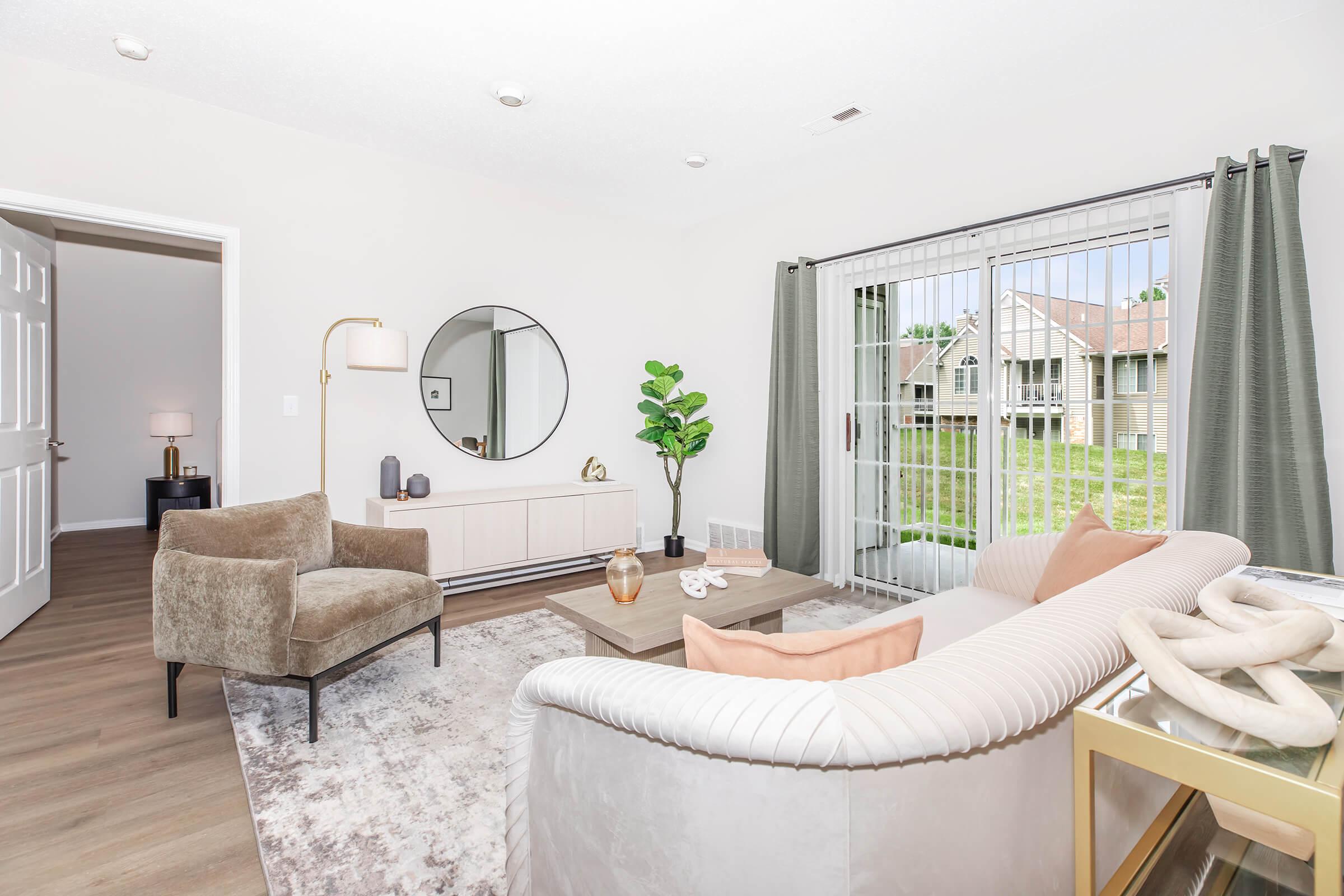 A modern living room featuring a light-colored sofa, a coffee table, and a plush armchair. Large windows with sheer curtains allow natural light. A decorative mirror and potted plant add a touch of elegance, while a neutral color palette creates a cozy atmosphere. The room opens to an outdoor view.