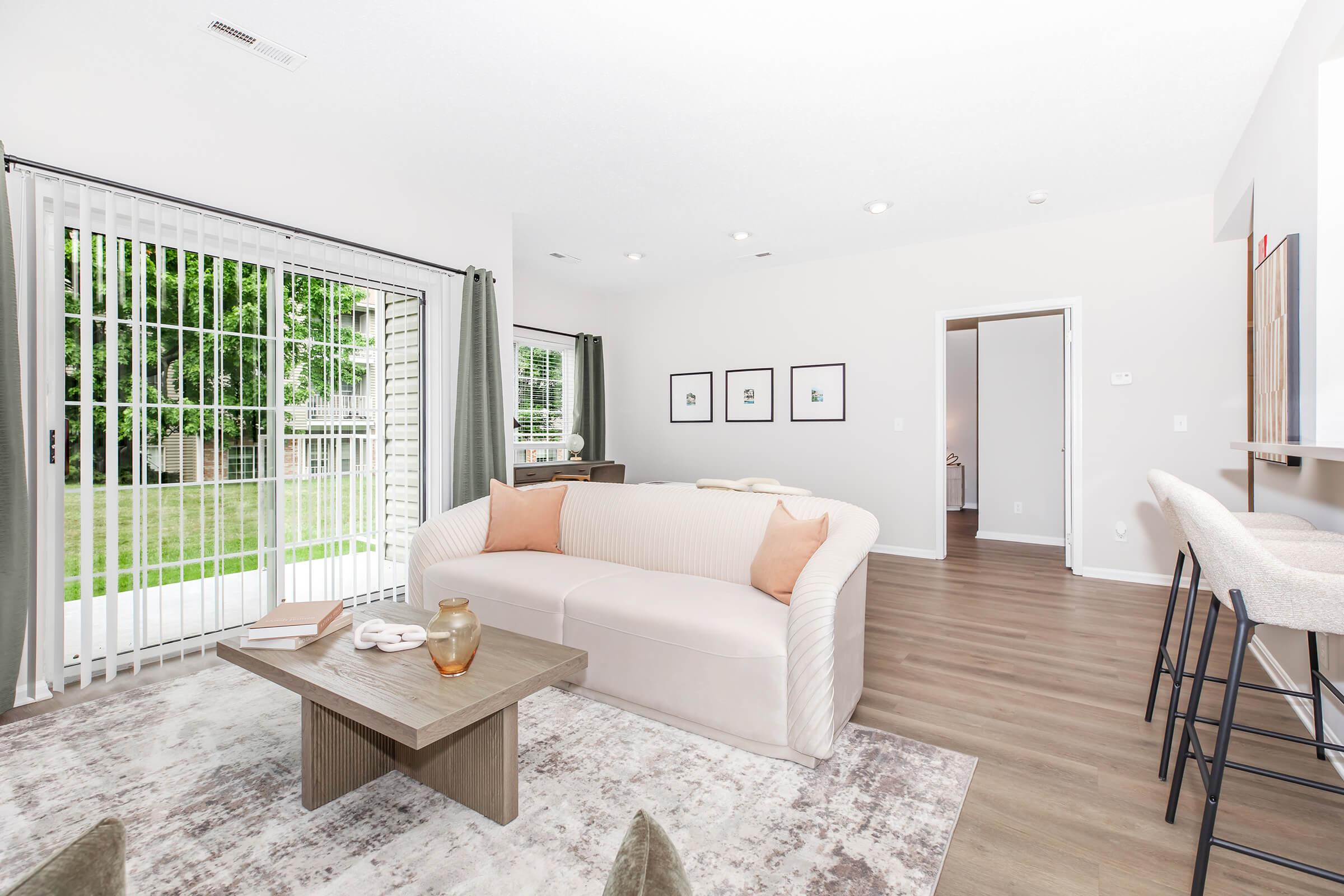 A modern living room featuring a light-colored sofa with cushions, a wooden coffee table, and a rug on a wooden floor. Large windows with sheer curtains let in natural light, and a set of framed pictures adorns the wall. A view of greenery is visible outside through the sliding glass door.