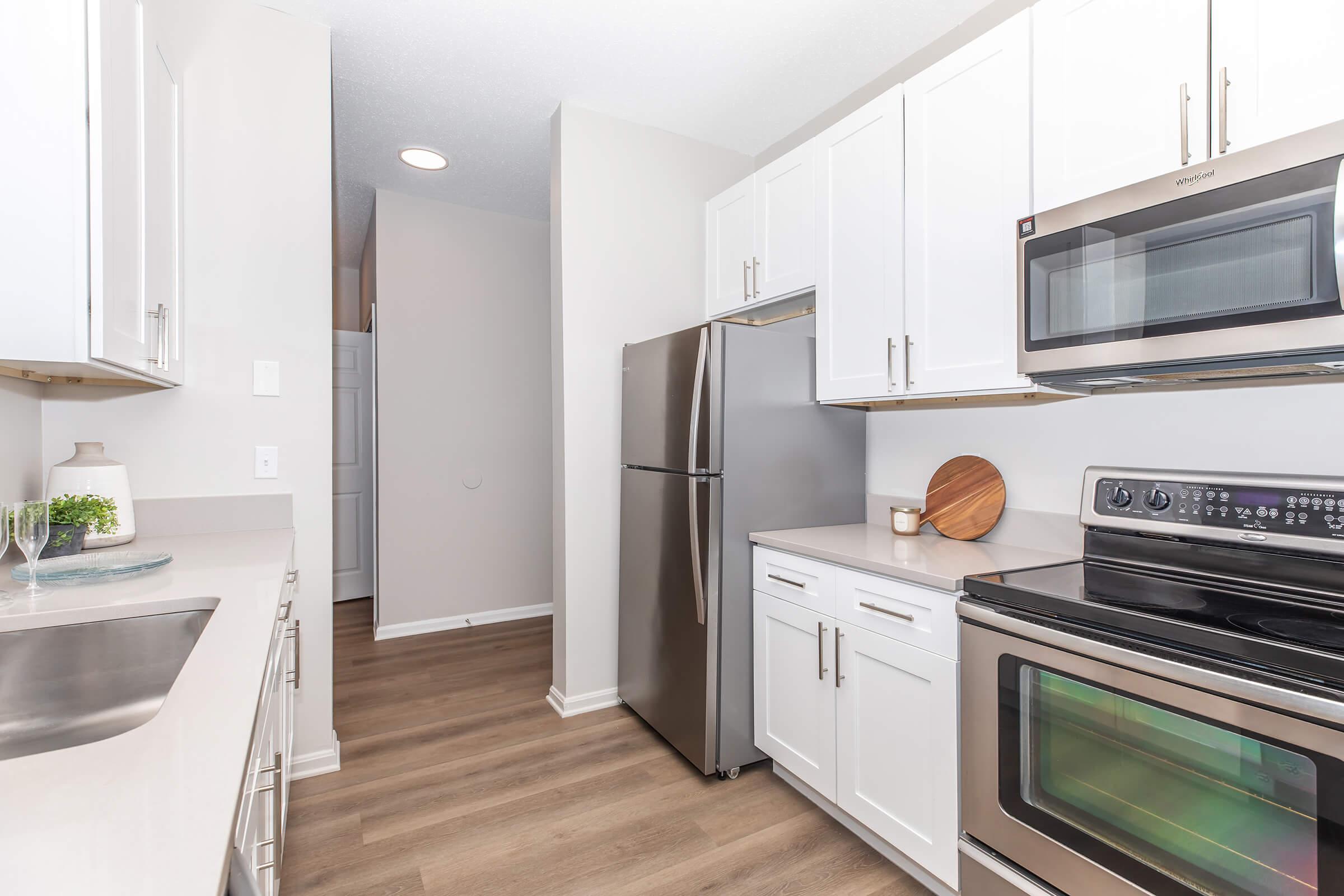 A bright and modern kitchen featuring stainless steel appliances, including a refrigerator and oven. The cabinetry is white with sleek handles, and a neutral countertop complements the wood-like flooring. A small wooden cutting board is placed on the countertop, adding a warm touch to the space.