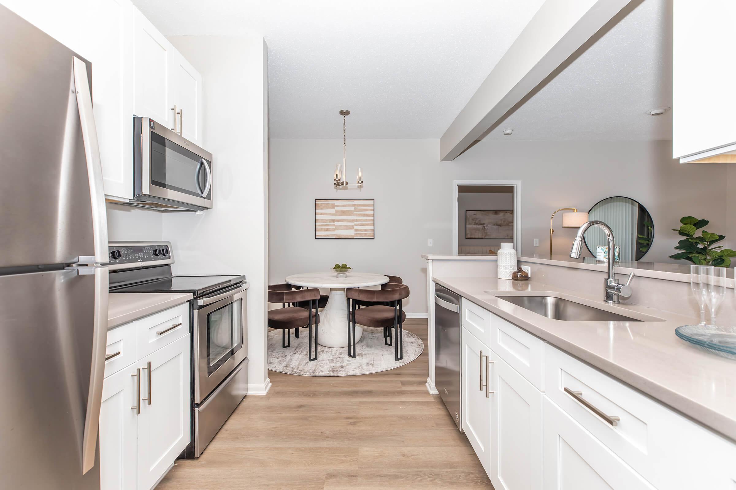 Modern kitchen featuring stainless steel appliances, including a refrigerator and oven. The sleek white cabinetry complements a light countertop. A dining area with a round table and four chairs is visible in the background, adorned with a circular rug and decorative elements that enhance the inviting atmosphere.