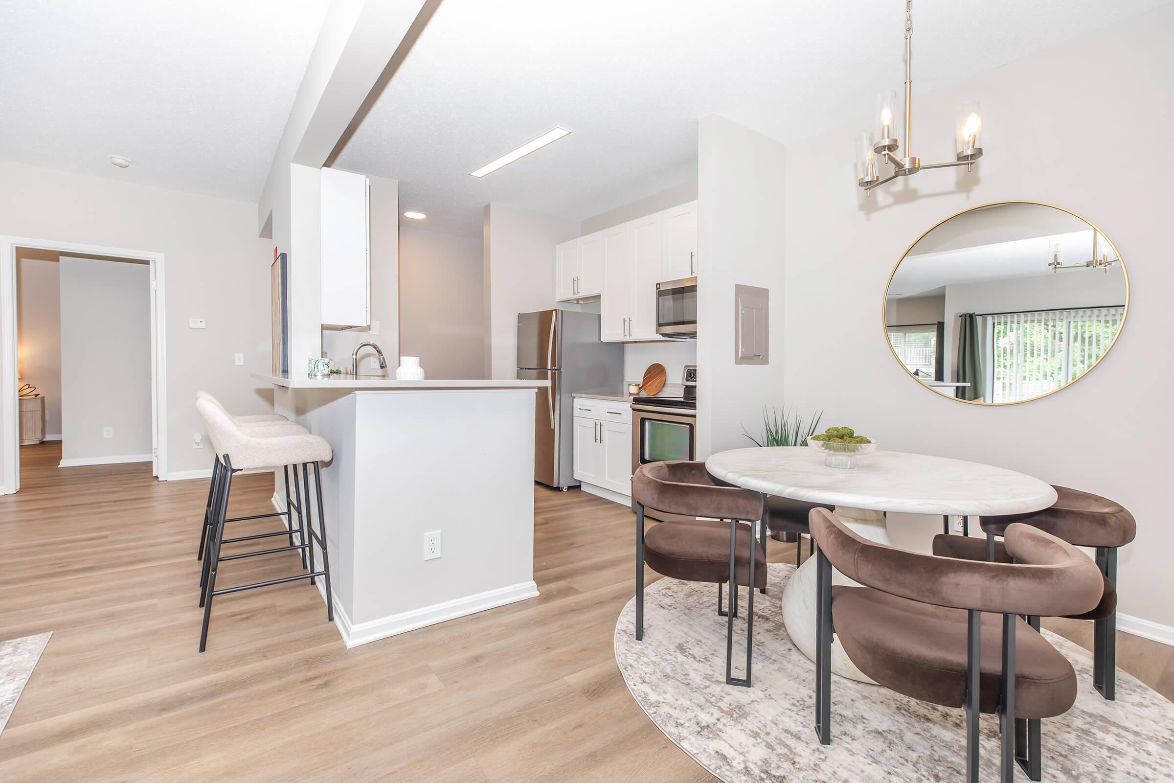 A modern kitchen and dining area featuring a round marble table surrounded by upholstered chairs, an open layout with light wood flooring, and a stylish kitchen with white cabinets and stainless steel appliances. Natural light flows in through large windows, enhancing the inviting atmosphere.