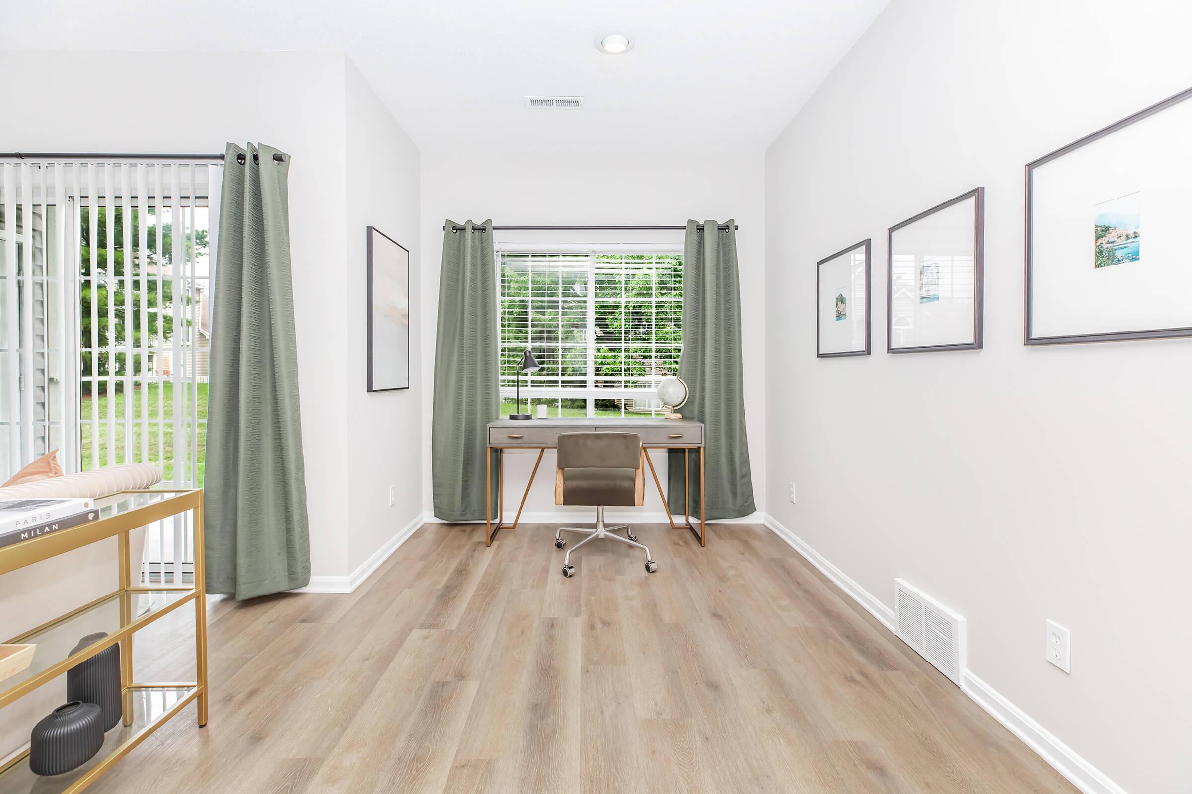 A brightly lit room with light wood flooring and neutral walls. It features a modern desk with a chair, framed artwork on the walls, and curtains covering a window that overlooks greenery. A small shelf holds decorative items, enhancing the workspace ambiance.