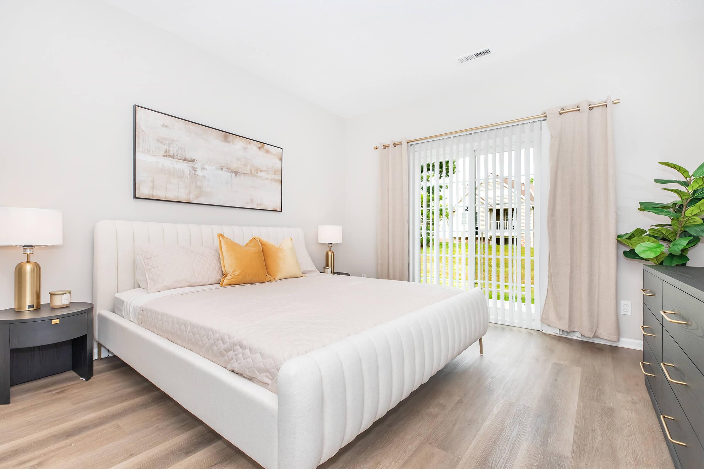 A modern bedroom featuring a sleek white upholstered bed with decorative pillows, a bedside table with a lamp on each side, and a dresser. The room has light-colored walls and large windows with vertical blinds, allowing natural light to fill the space. A green plant adds a touch of nature.