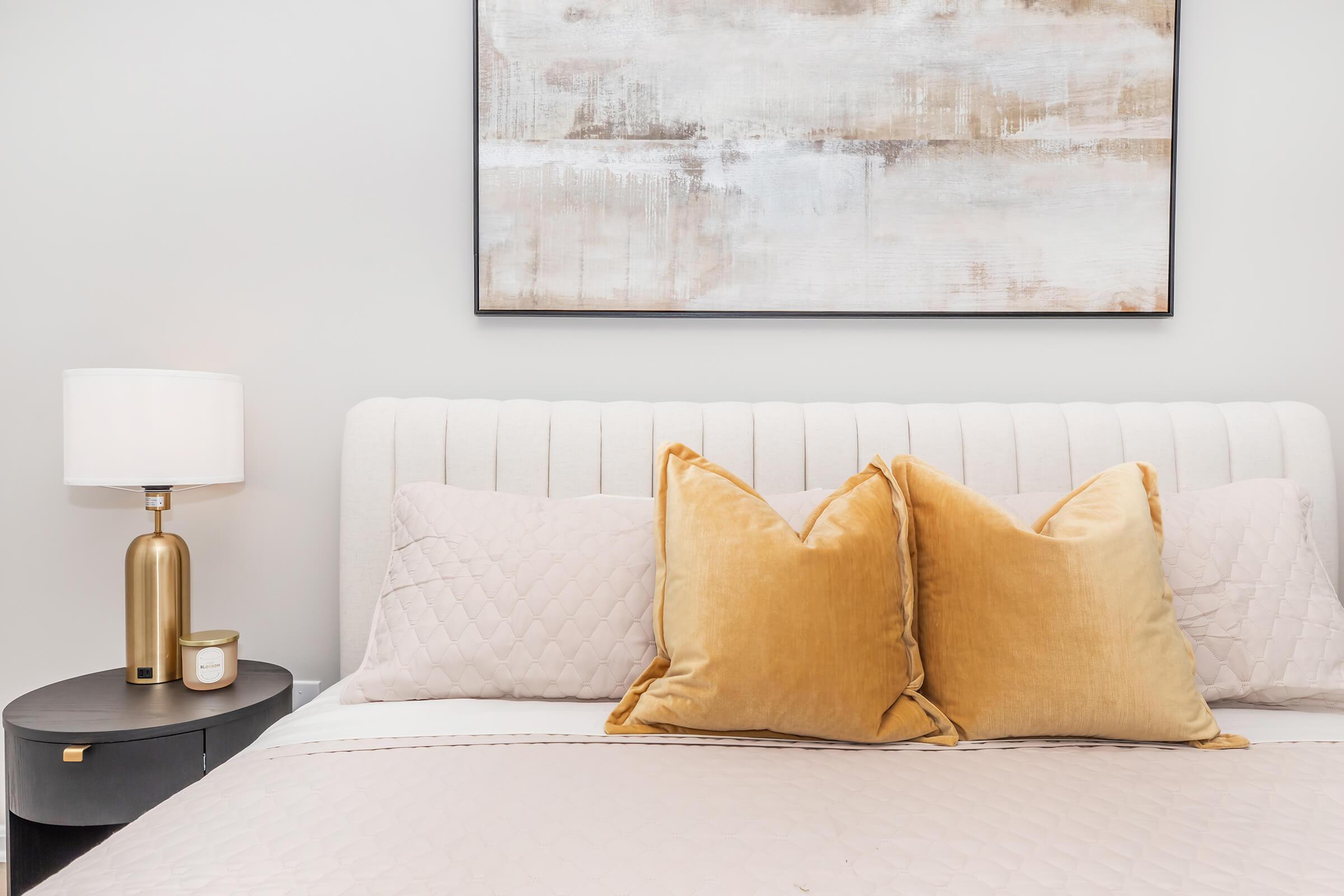 A neatly made bed with a light-colored headboard featuring two large, golden velvet pillows. A cozy beige quilt covers the bed. Next to it, a round nightstand holds a stylish lamp with a white shade and a small candle, accompanied by a modern abstract artwork on the wall above.