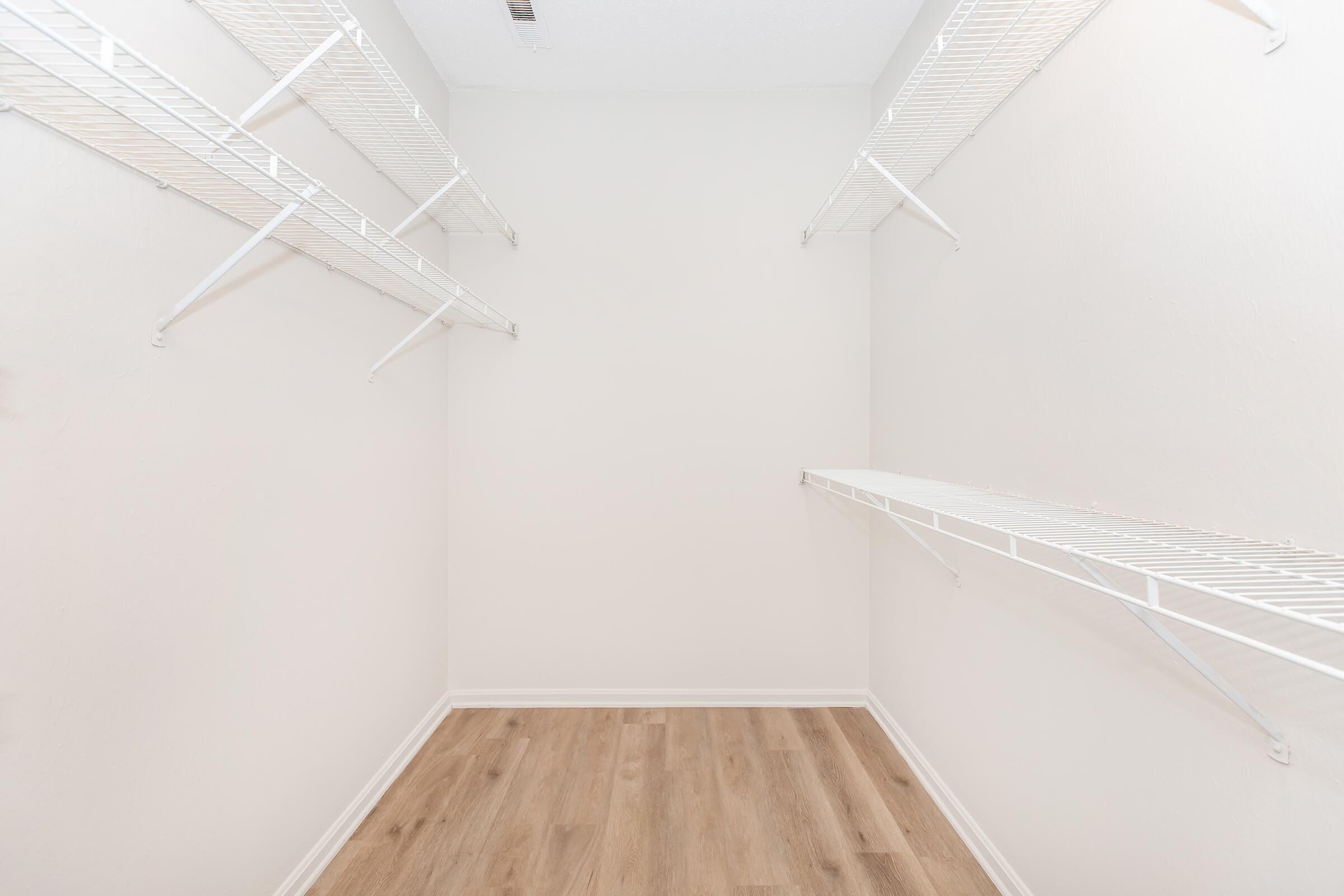 Empty walk-in closet featuring white wire shelves mounted on the walls. The walls are painted a light color, and the floor is a light wooden laminate. The space is well-lit, creating a clean and open feel.