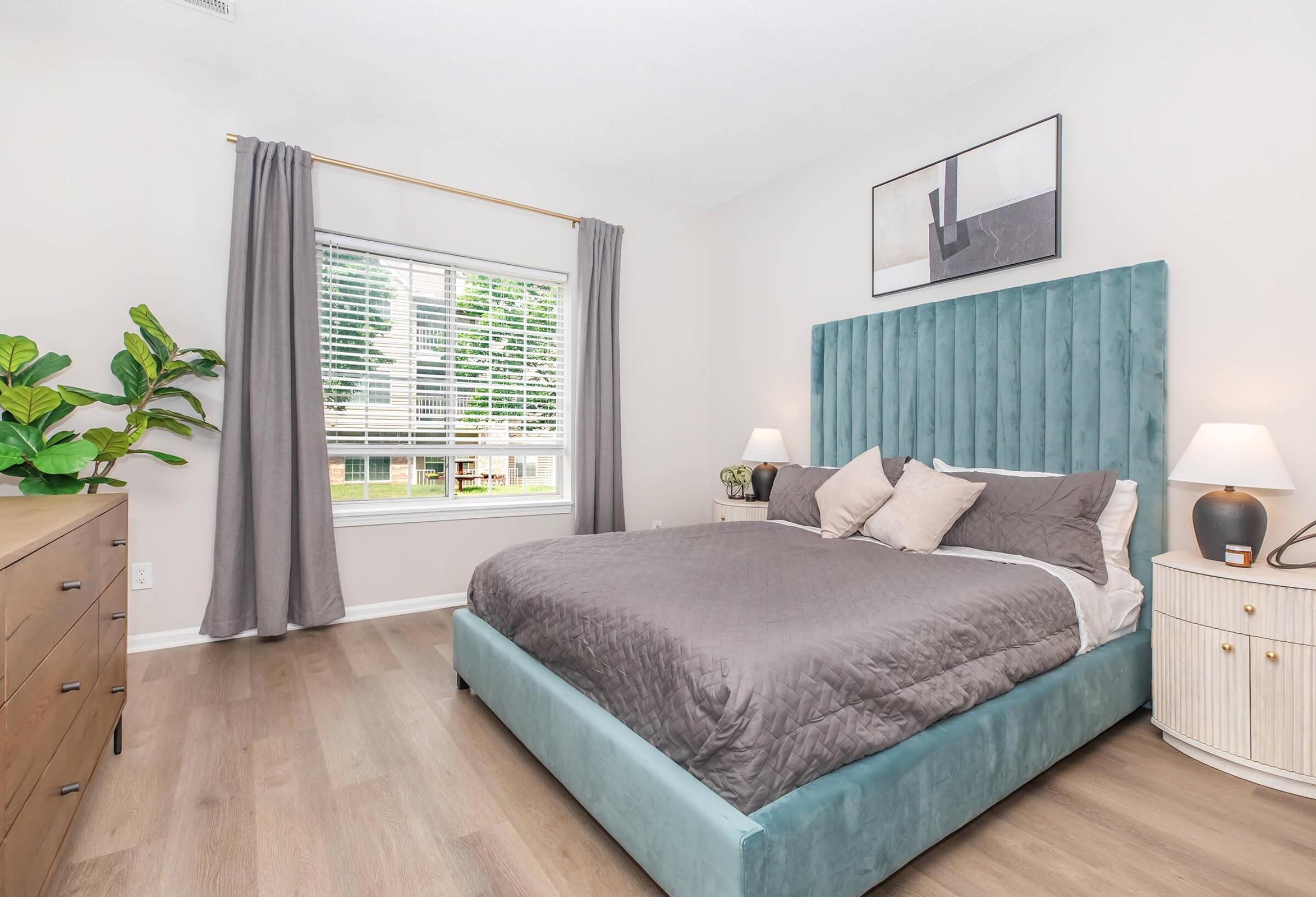 A modern bedroom featuring a light blue upholstered bed with gray bedding. There are two bedside lamps on either side, a wooden dresser, and a large window with sheer curtains letting in natural light. Decorative plants and minimalist wall art complete the stylish decor.