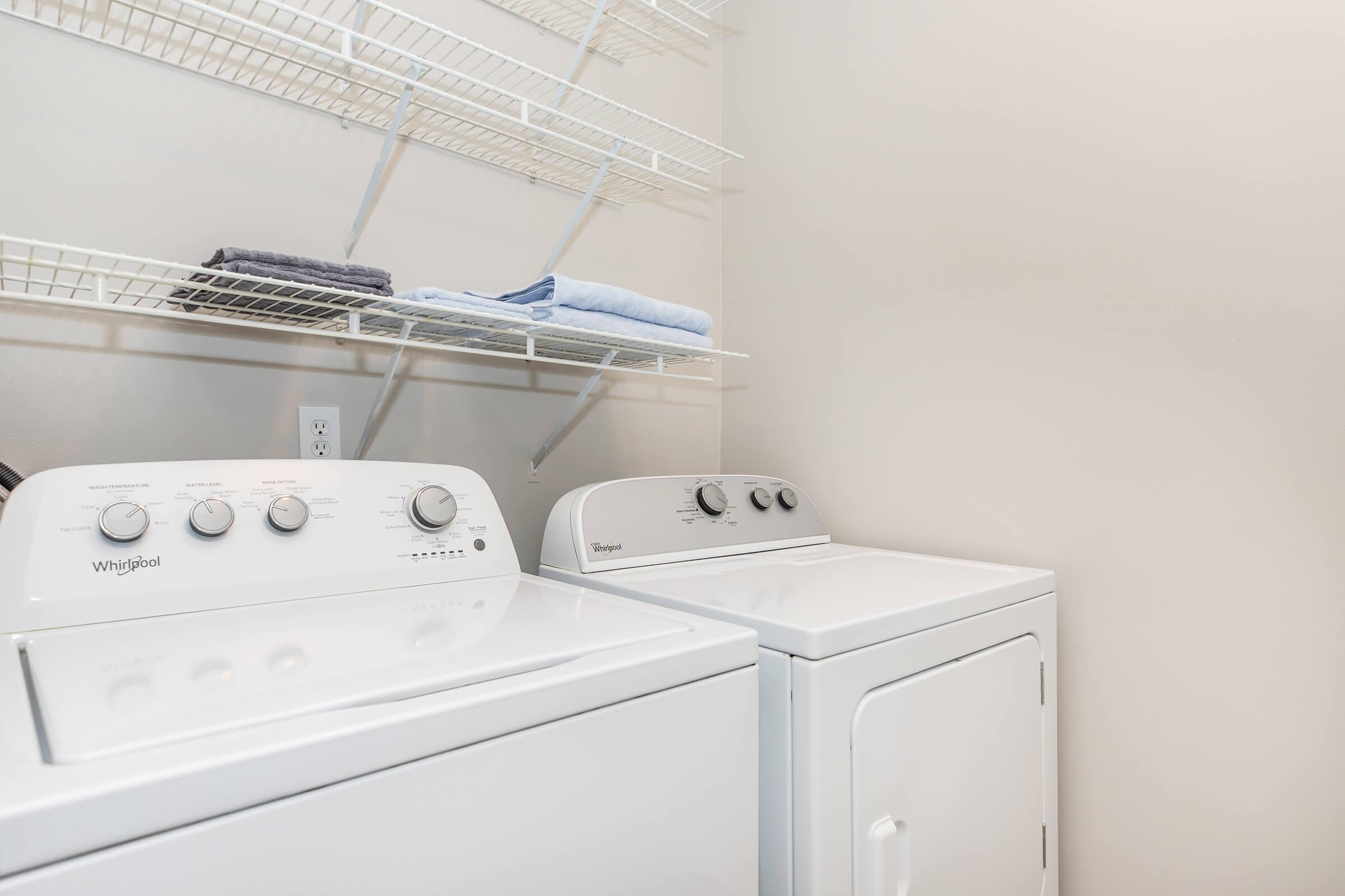 A clean laundry room featuring a white washer and dryer side by side. Above, there is a wire shelf holding neatly folded towels. The walls are a light neutral color, creating a bright and organized space for doing laundry.