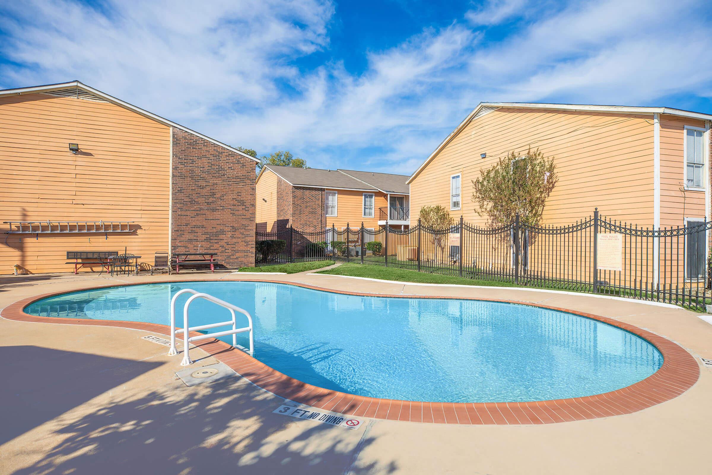 A clear blue swimming pool surrounded by a tan-colored, fenced area, with two neighboring buildings in the background. The sky is partly cloudy, creating a bright and inviting atmosphere, ideal for relaxation or leisure.