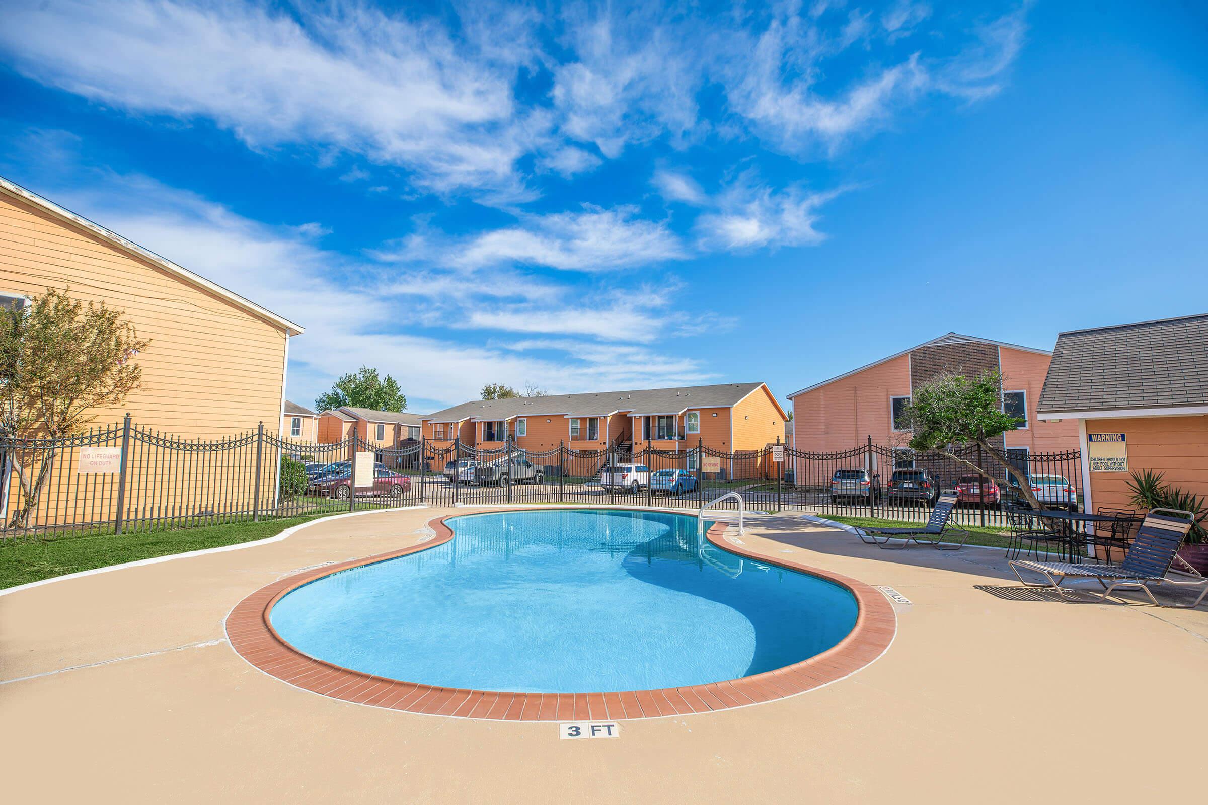 A clear blue swimming pool surrounded by a concrete deck, with lounge chairs nearby. In the background, there are two-story orange apartment buildings and a sunny, blue sky with a few clouds. A fenced area provides privacy to the pool.