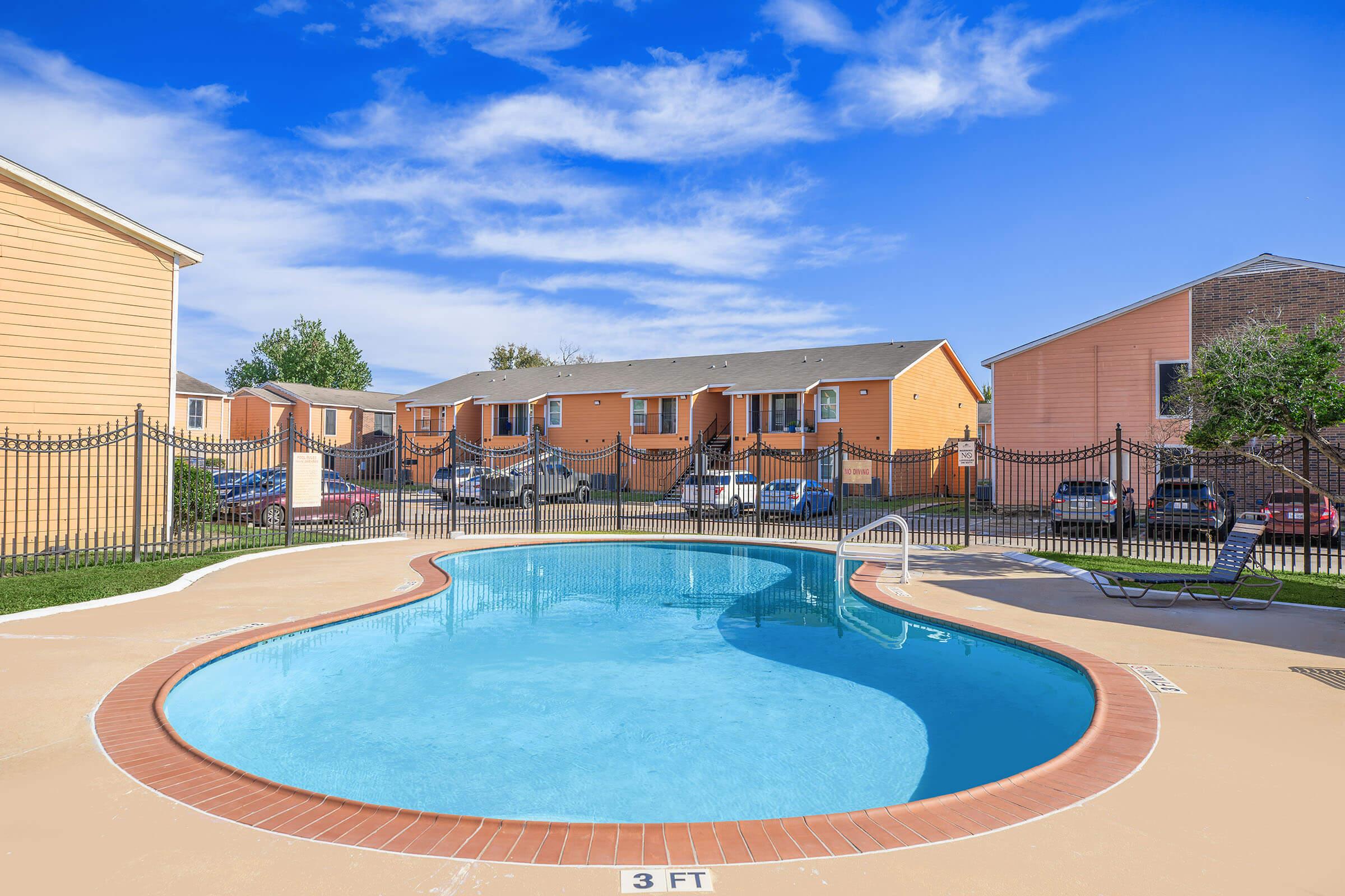 A clear blue swimming pool in a residential area, surrounded by a fence. In the background, there are several orange and beige apartment buildings with cars parked nearby under a bright blue sky with some clouds.