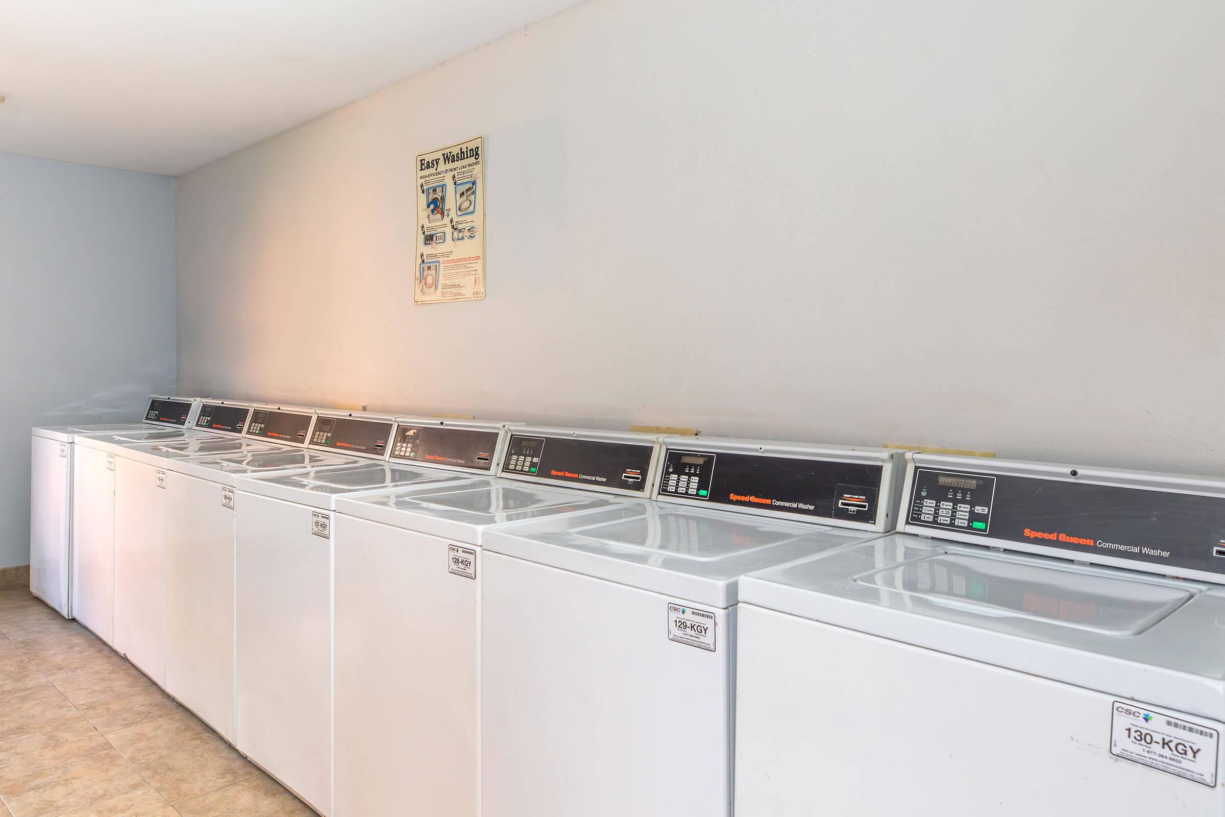 A row of seven white top-loading washing machines in a laundry room. The machines have black control panels with various settings. The walls are a light gray, and a framed notice is mounted on the wall above the machines. The floor is tiled in beige. The space is well-lit and tidy.