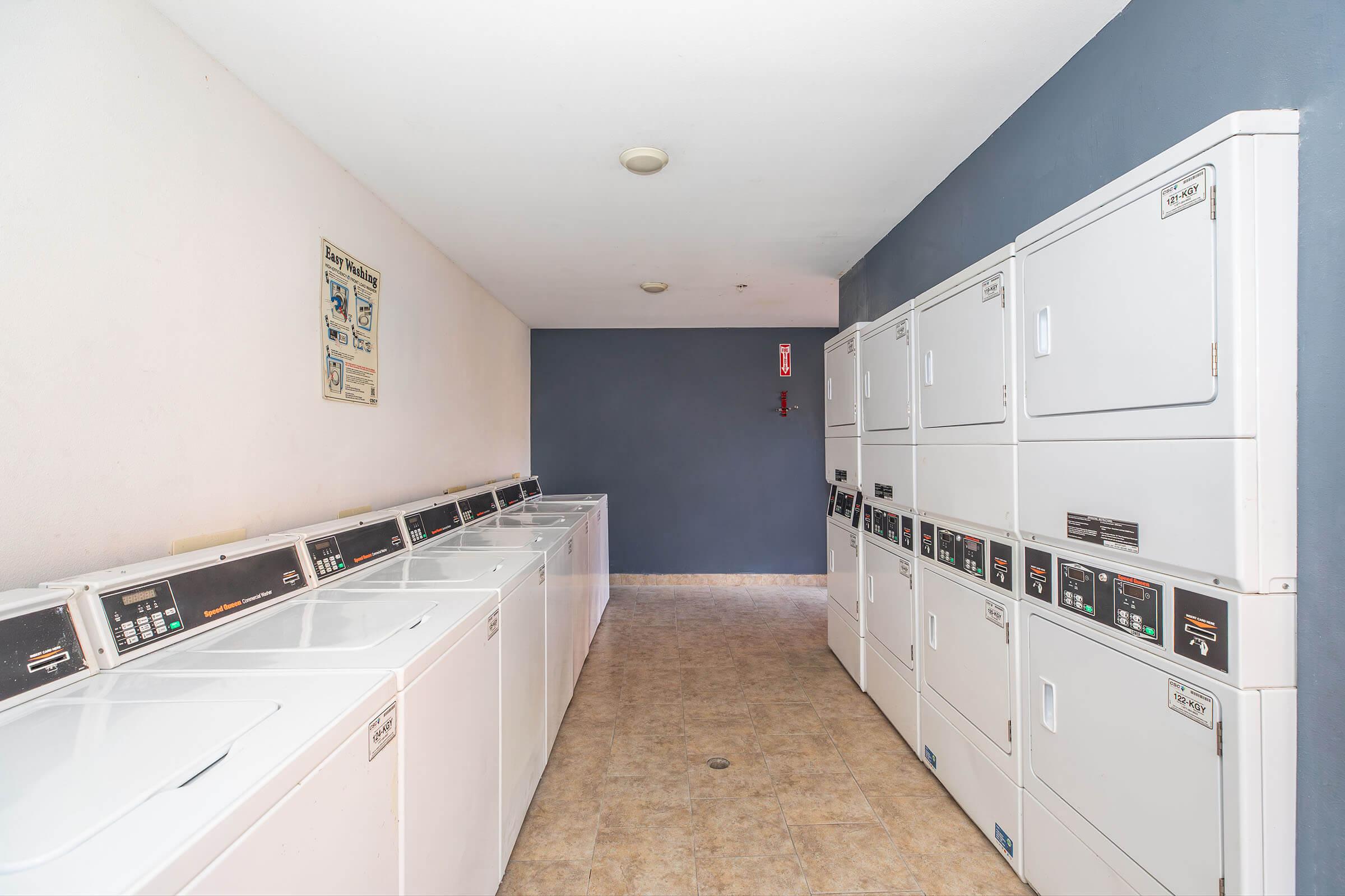 A clean and well-lit laundry room featuring multiple white washing machines and dryers lined up against the wall. The floor is tiled, and a simple blue wall provides the background. There is also a notice board on the left side with informational posters.