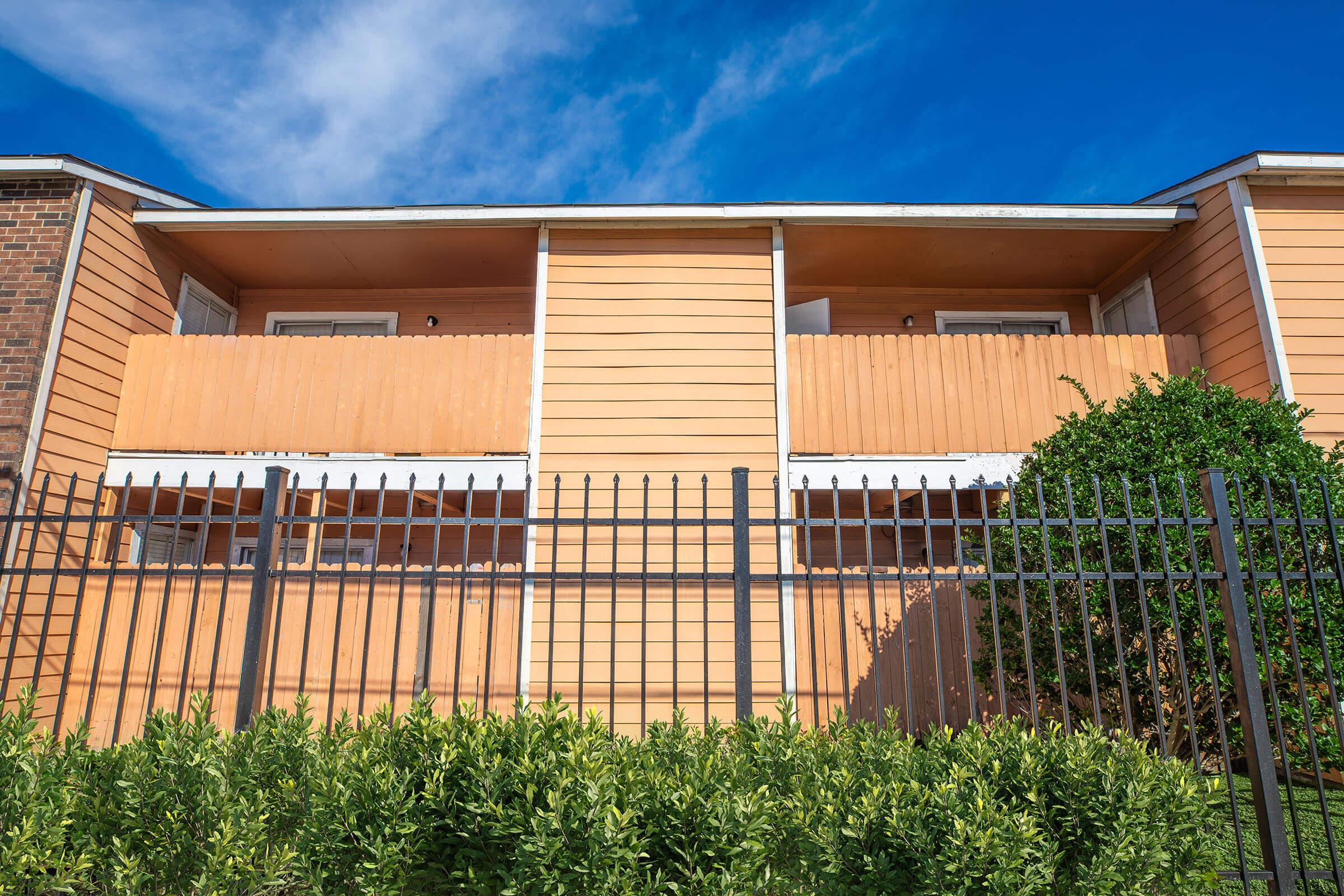 Exterior view of an orange apartment building with a balcony on the second floor. The building is surrounded by a black fence and green shrubs at the base. A clear blue sky with a few clouds is visible above.